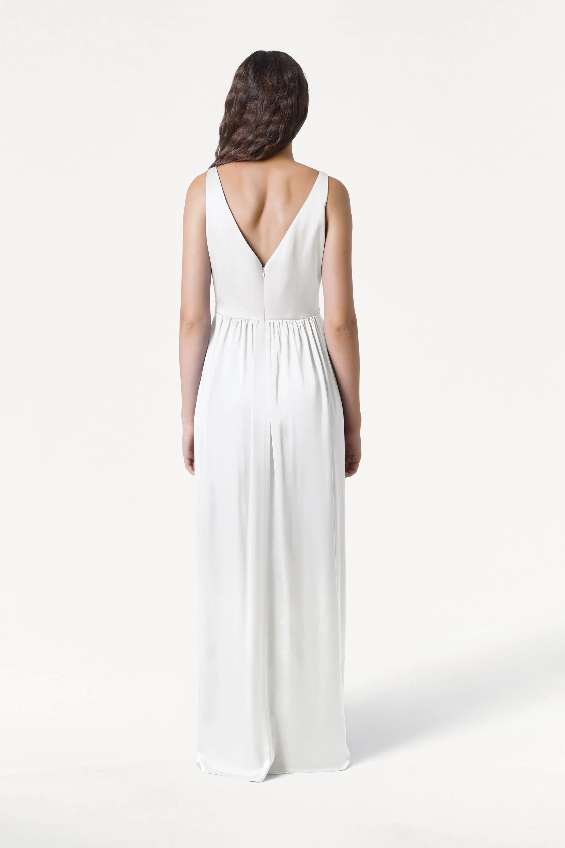 A woman with long, wavy brown hair faces away in the Carmen Dress in Ivory Satin by TH&TH—a sleeveless, floor-length gown with a deep V-back and gathered waist—ideal for modern bridal parties, set against a plain white background.