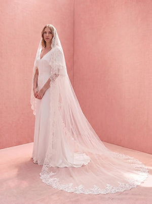 Woman wearing a white lace wedding dress with a long veil against a pink background