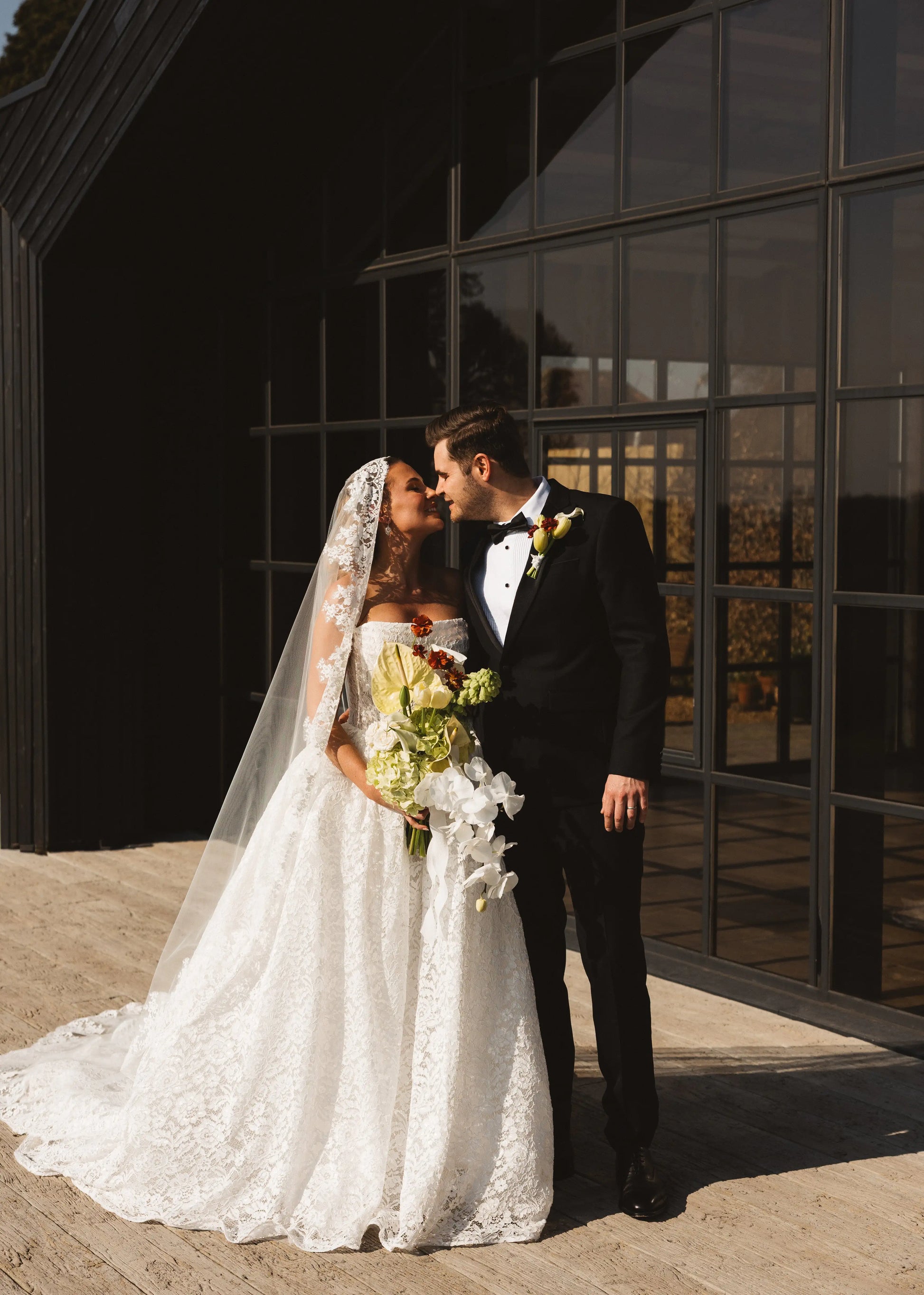 A bride in a white lace gown and TH&TH's Catherine Eyelash Lace Mantilla Veil smiles, holding a bouquet as she embraces a groom in a black tuxedo outside a modern building with large glass windows.