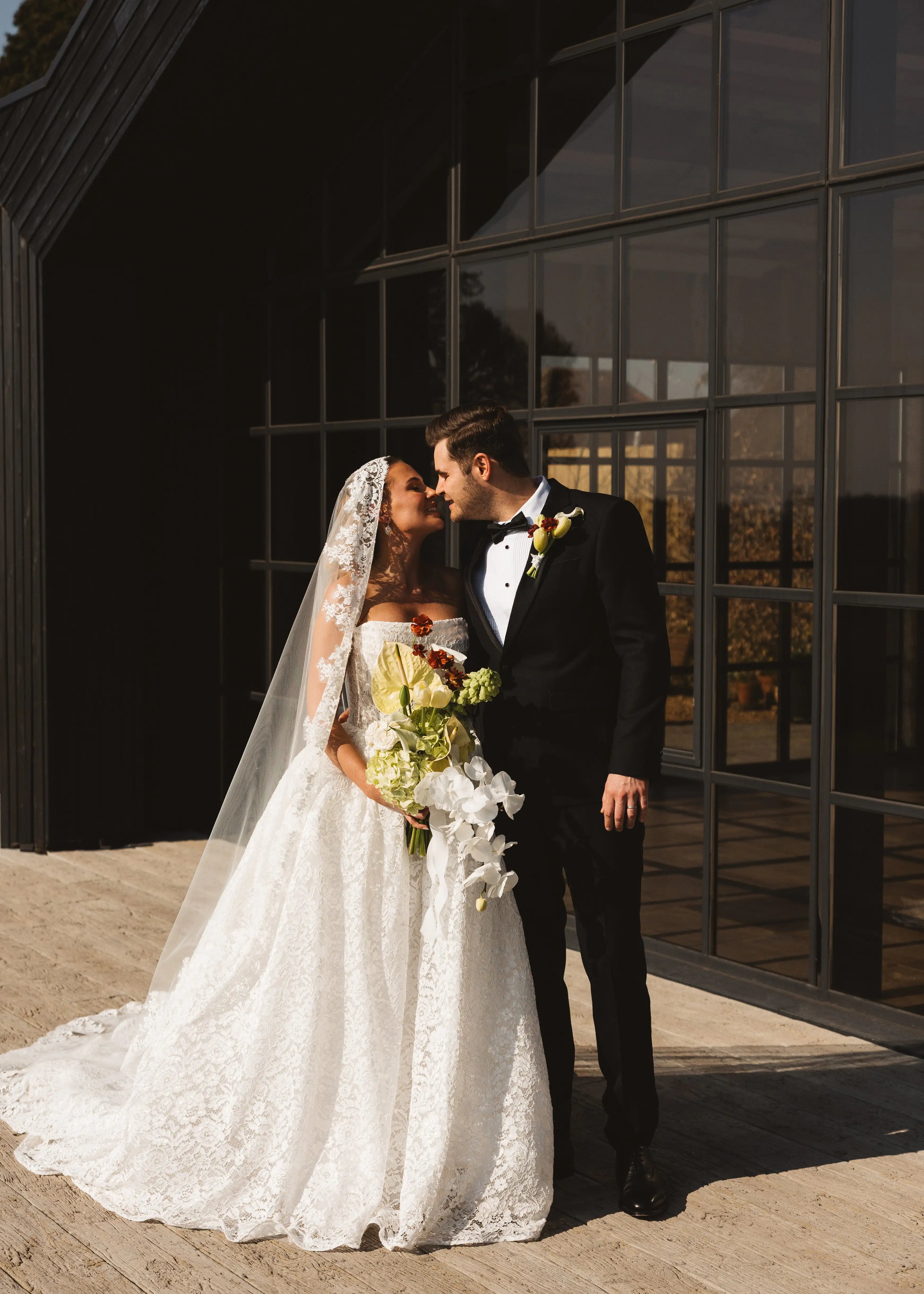 A bride in a white lace gown and TH&TH's Catherine Eyelash Lace Mantilla Veil smiles, holding a bouquet as she embraces a groom in a black tuxedo outside a modern building with large glass windows.