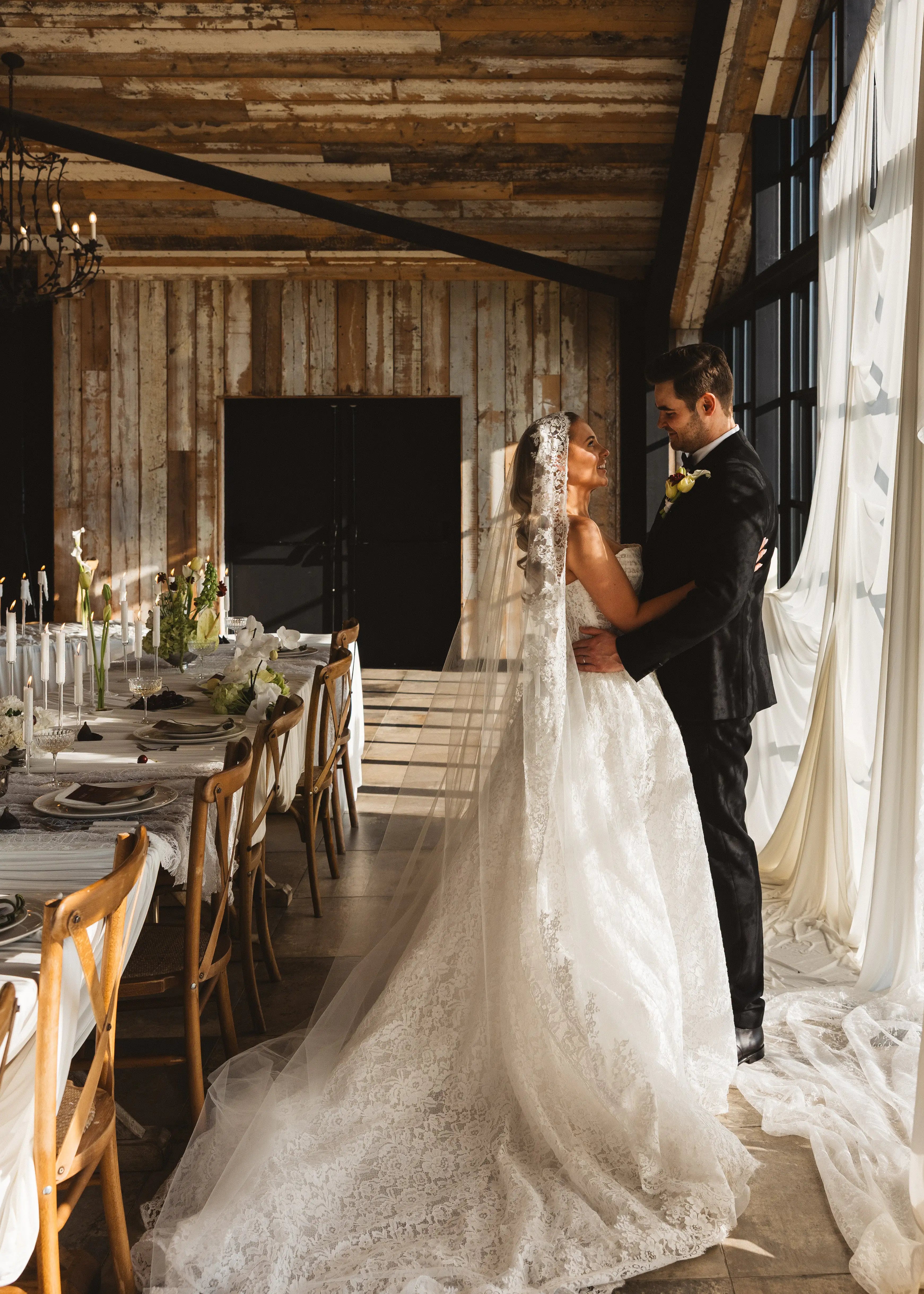 Wearing the Catherine Eyelash Lace Mantilla Veil by TH&TH, a bride stands with her groom in a sunlit rustic room. They share a loving gaze beside a decorated dining table, capturing an intimate wedding moment.