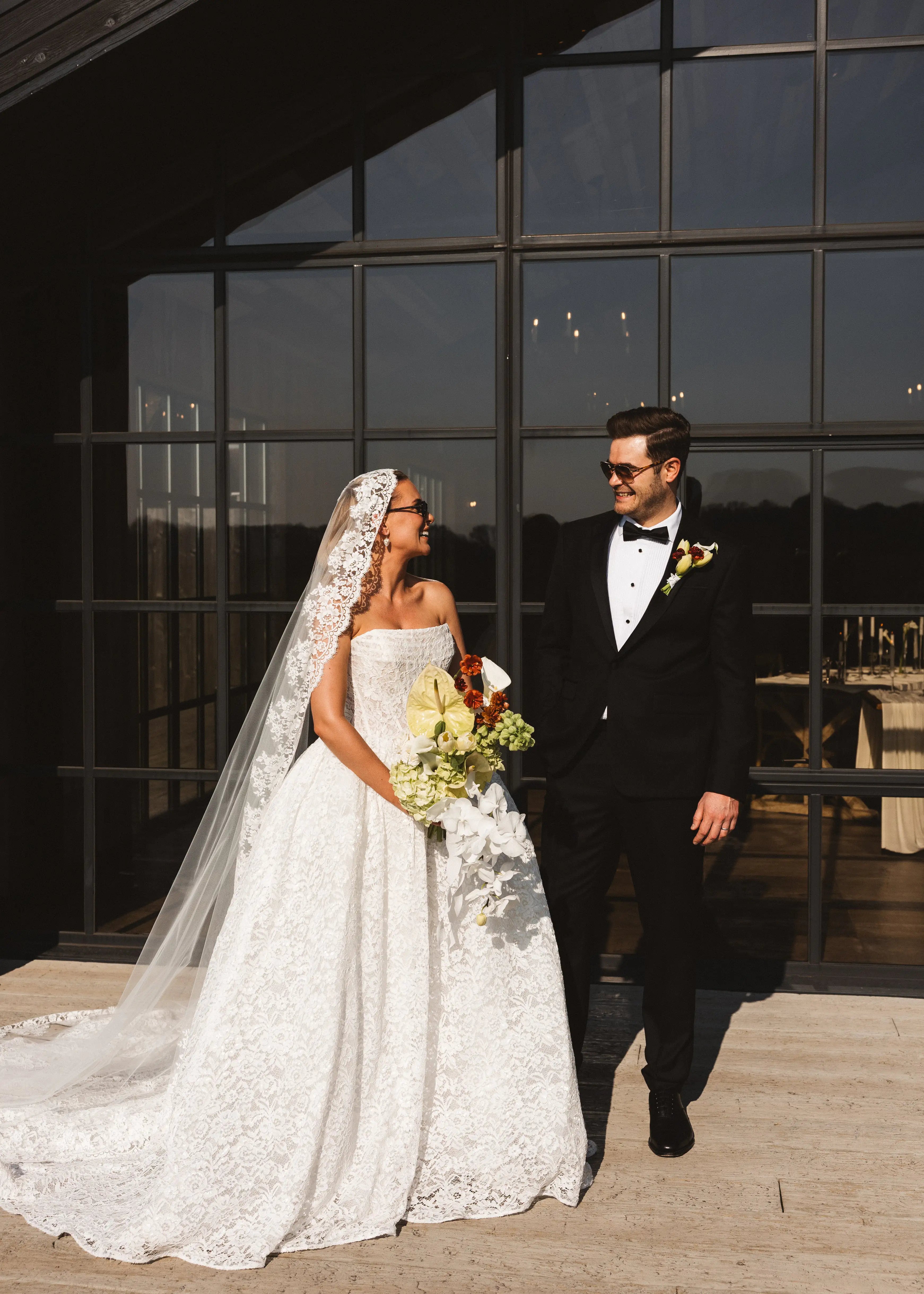 A bride in a white gown and the Catherine Eyelash Lace Mantilla Veil by TH&TH holds a bouquet beside her groom in a black tuxedo. They smile at each other outside a modern building with large glass windows.