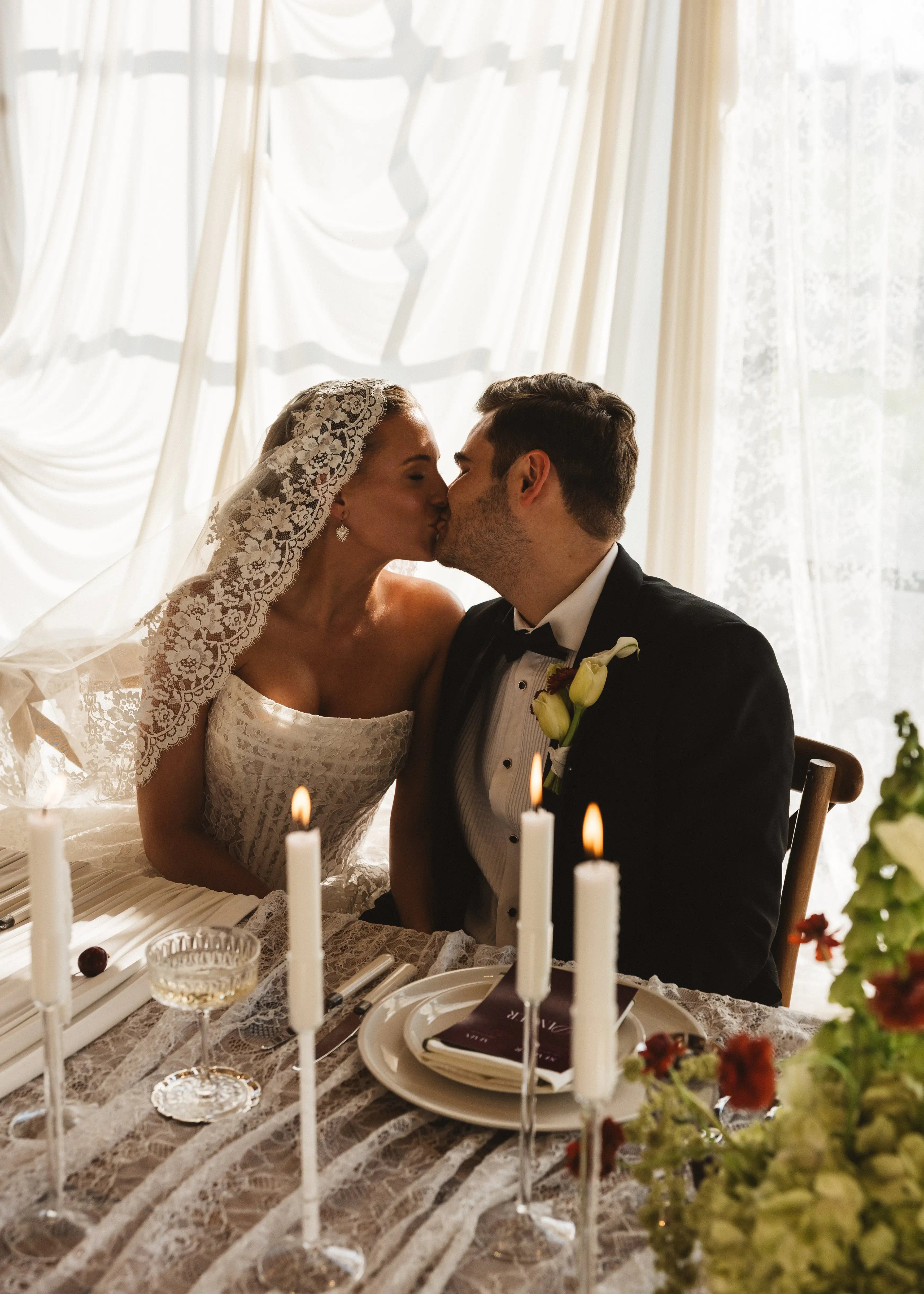 A bride in the Catherine Eyelash Lace Mantilla Veil by TH&TH shares a kiss with her groom at their candlelit, flower-adorned wedding table, as soft sunlight streams through sheer curtains behind them.