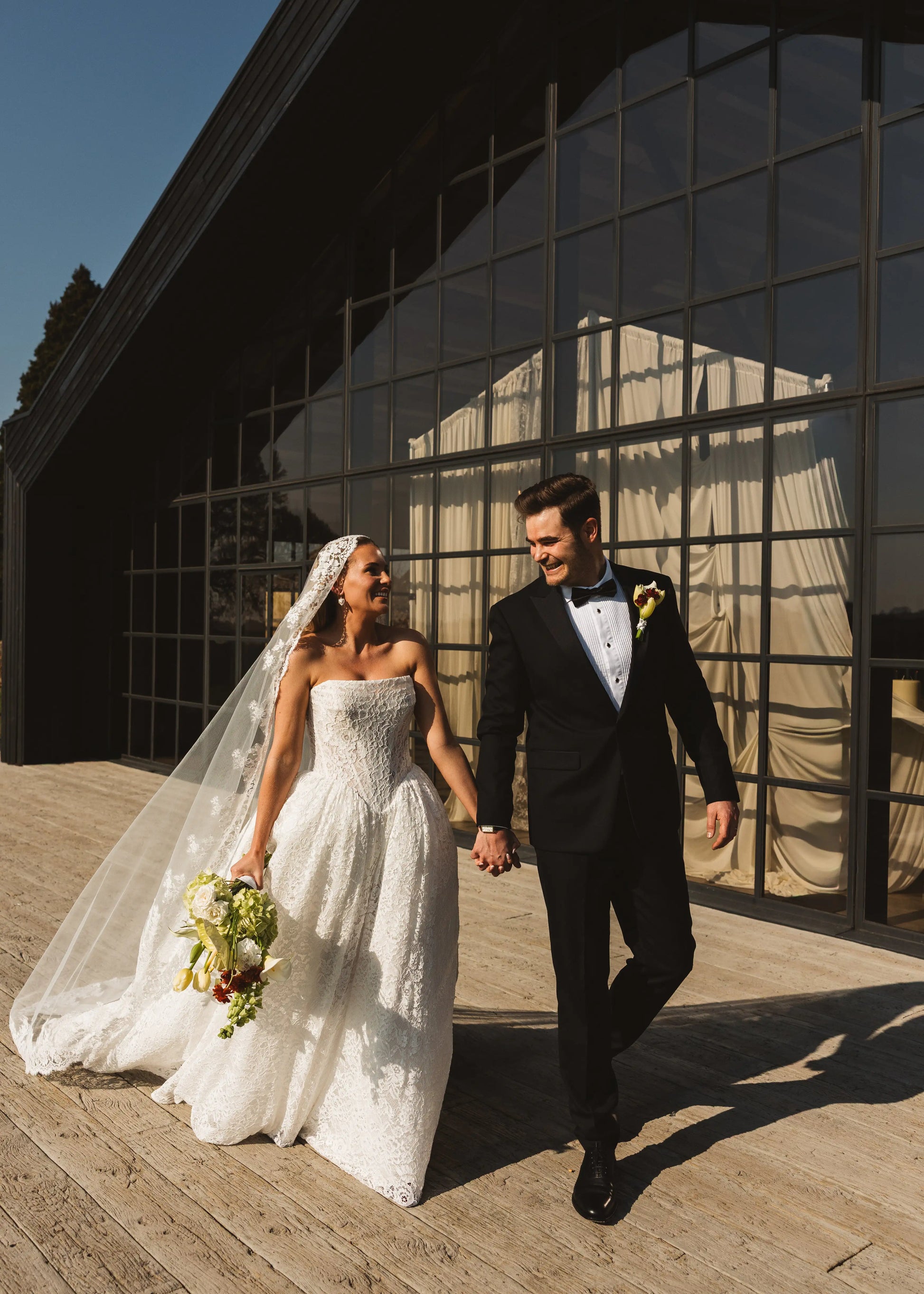 A bride in a white dress and the Catherine Eyelash Lace Mantilla Veil by TH&TH holds a bouquet and smiles at her groom in a black tuxedo as they walk hand in hand outside a modern glass building on a sunny day.