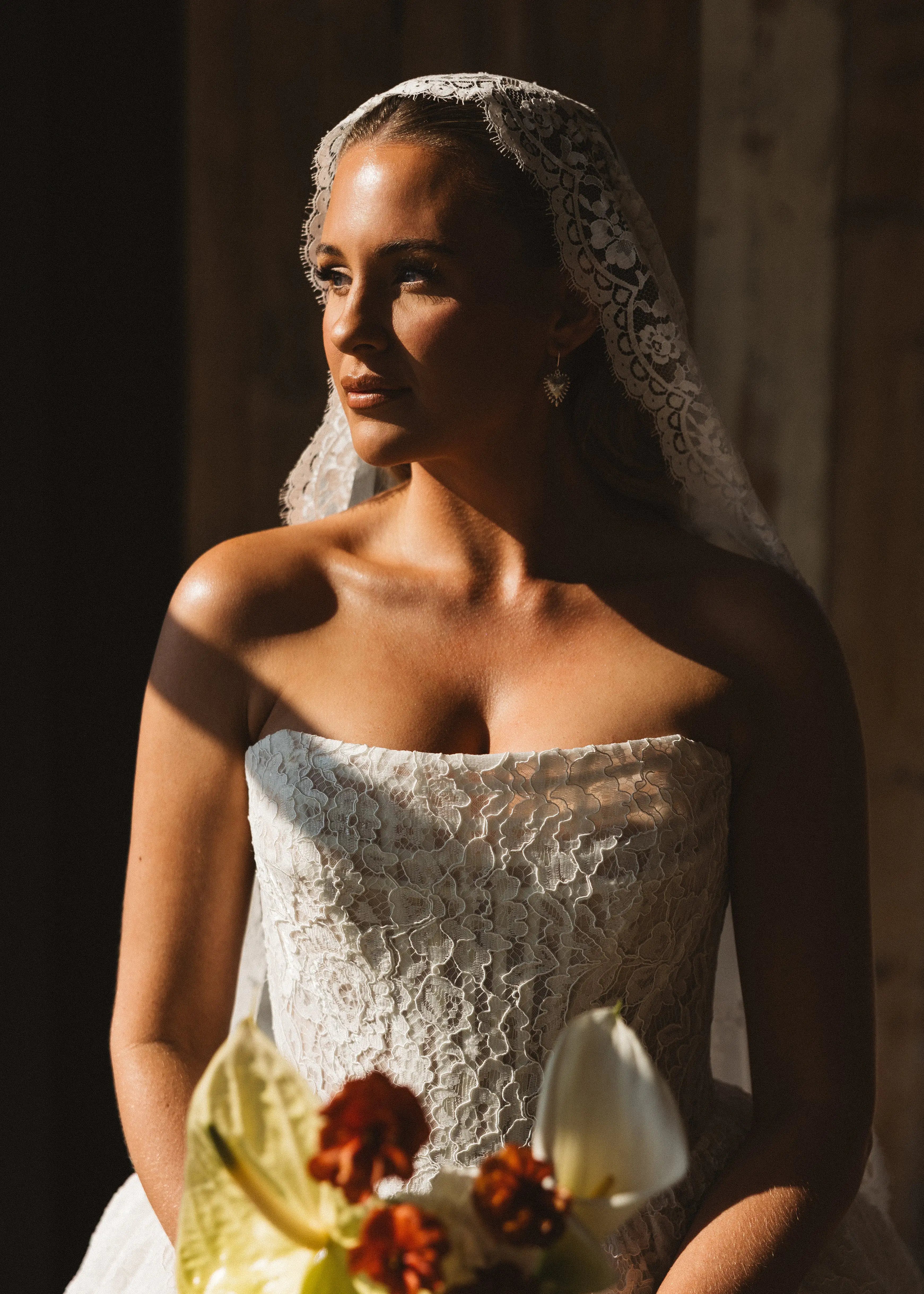 A bride wearing the TH&TH Catherine Eyelash Lace Mantilla Veil sits in sunlight with a bouquet of white and orange flowers, gazing thoughtfully to the side.