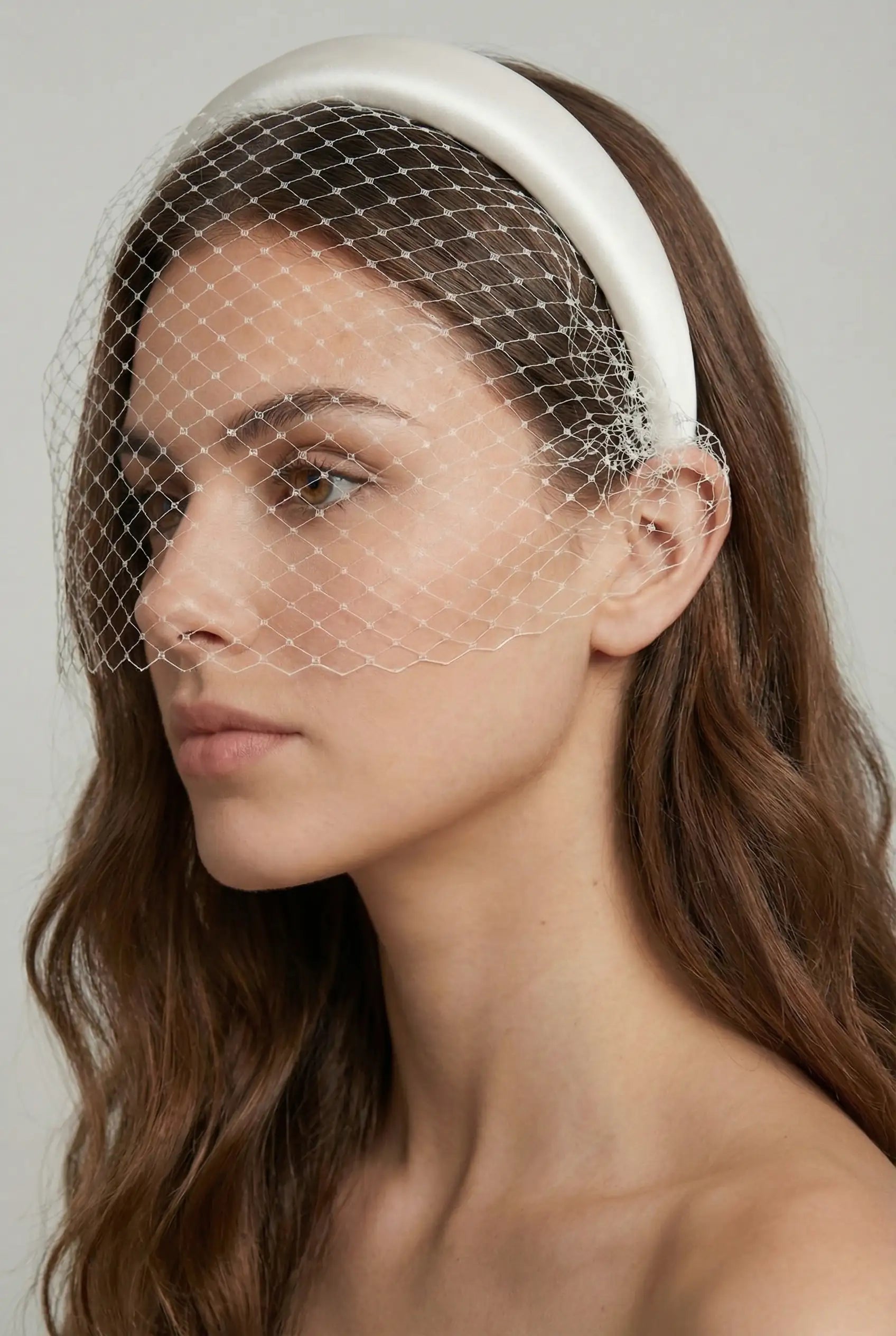 A woman with long brown hair wears the TH&TH Cavendish Headband with Birdcage veil, posing against a plain background.