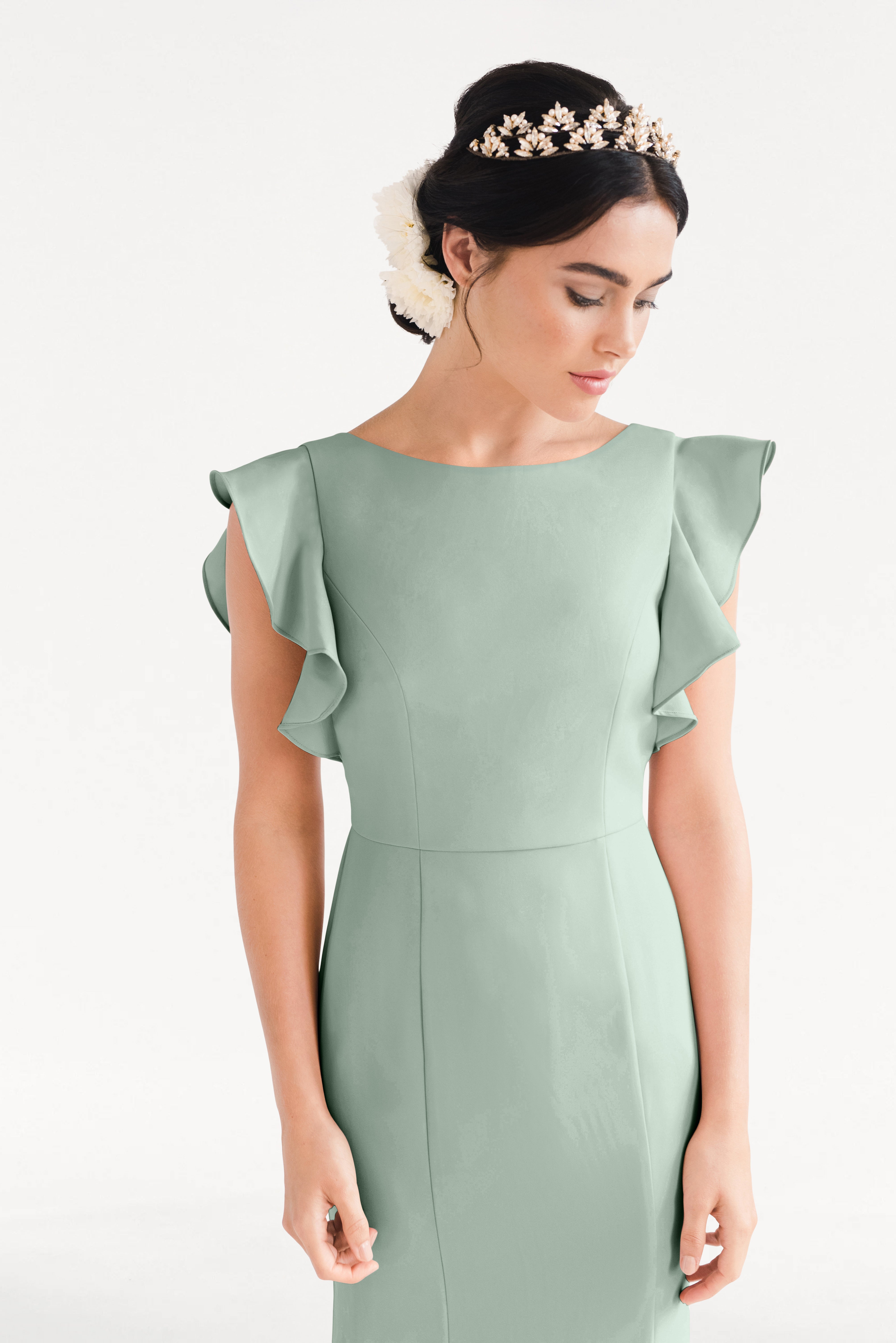 A woman in the TH&TH Cecelia Moss Crepe Bridesmaid Dress in Sage Green with ruffled sleeves and a floral headpiece stands against a plain white background, looking down with her hands by her sides.