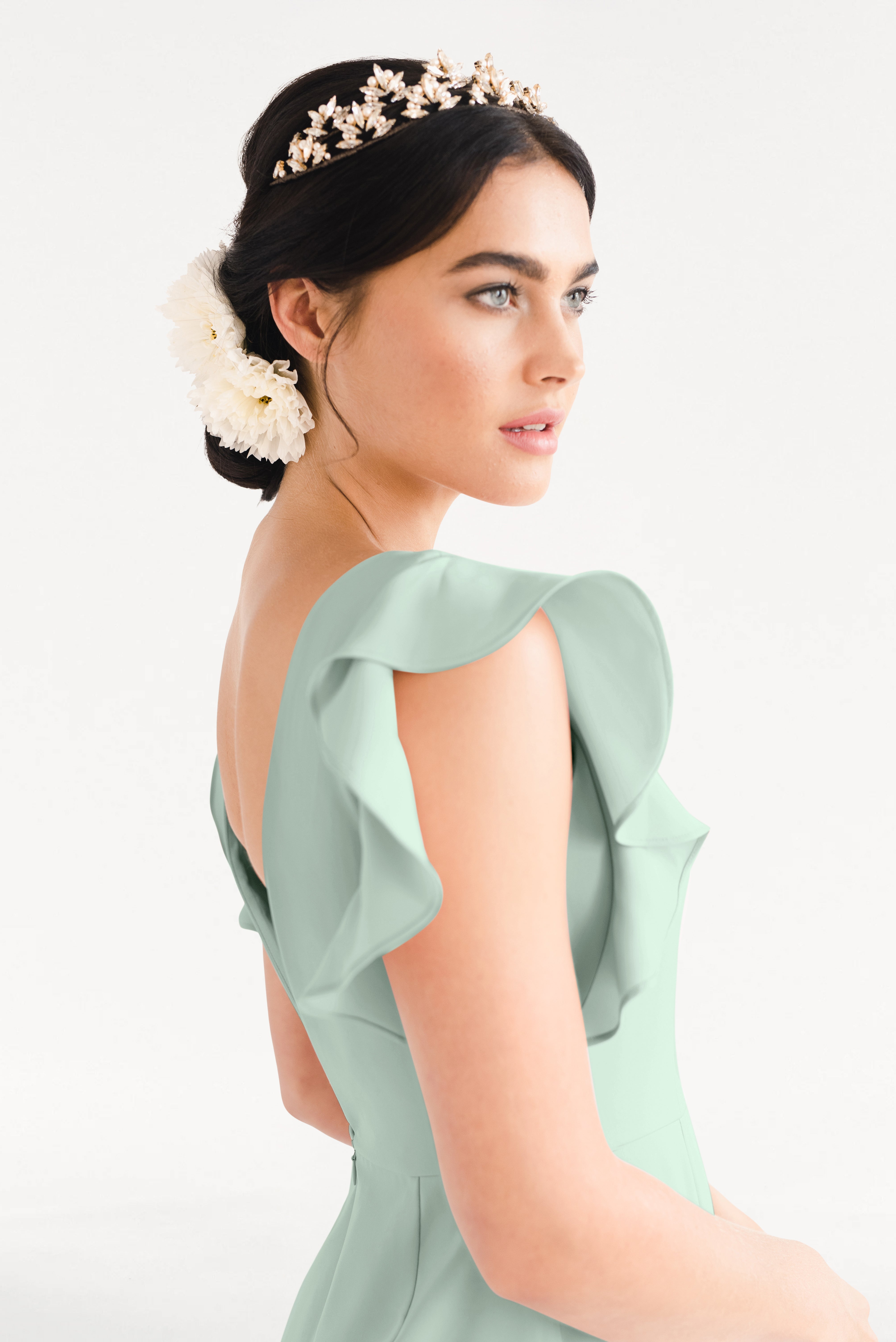 A woman with dark hair in a low bun, adorned with white flowers and a gold floral headband, wears the TH&TH Cecelia Moss Crepe Bridesmaid Dress in Sage Green with ruffled sleeves, standing against a plain white background.