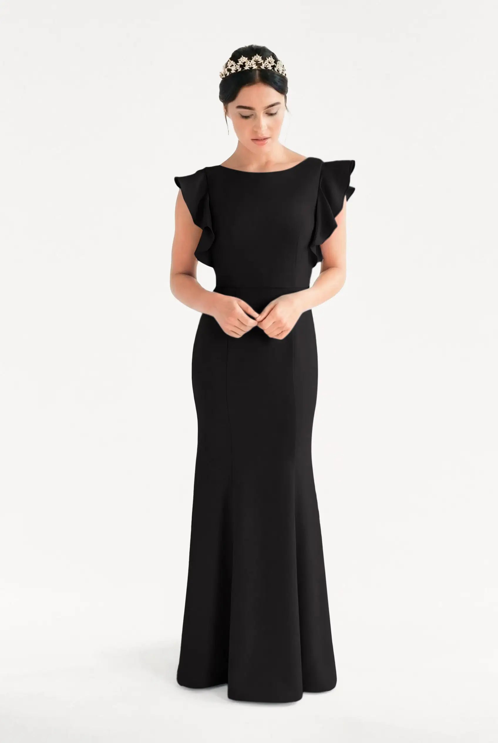 A woman in the TH&TH Cecelia Moss Crepe Bridesmaid Dress in Black, featuring ruffled shoulders, stands against a white background. She wears a jeweled headband, looks down, and holds her hands together in front of her.