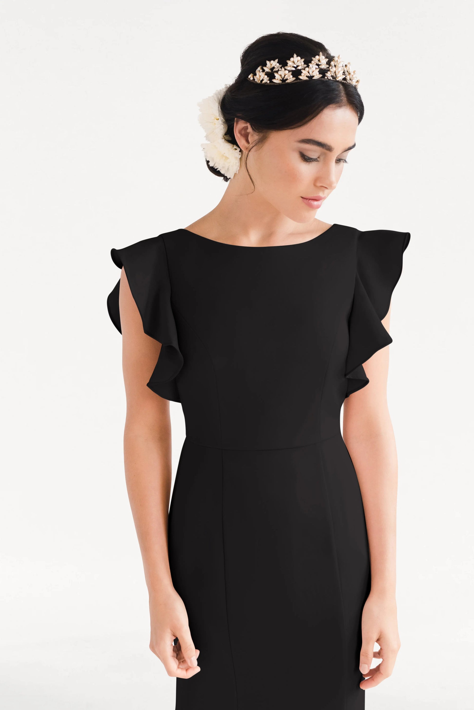 A woman in the Cecelia Moss Crepe Bridesmaid Dress in Black by TH&TH, featuring ruffled sleeves and a floral headpiece, looks down against a plain white background.