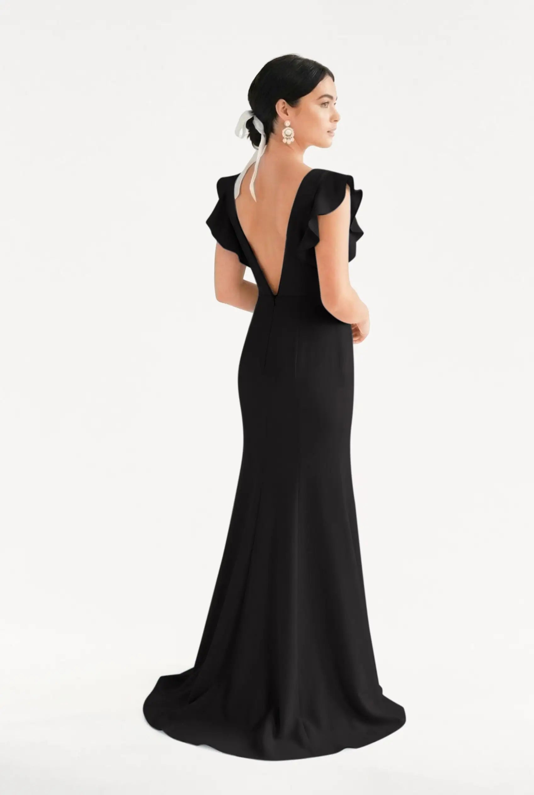 A woman stands facing away in the Cecelia Moss Crepe Bridesmaid Dress in Black by TH&TH, featuring ruffled sleeves and a deep V-back. Her dark hair is tied with a white ribbon, statement earrings visible, set against a plain white background.