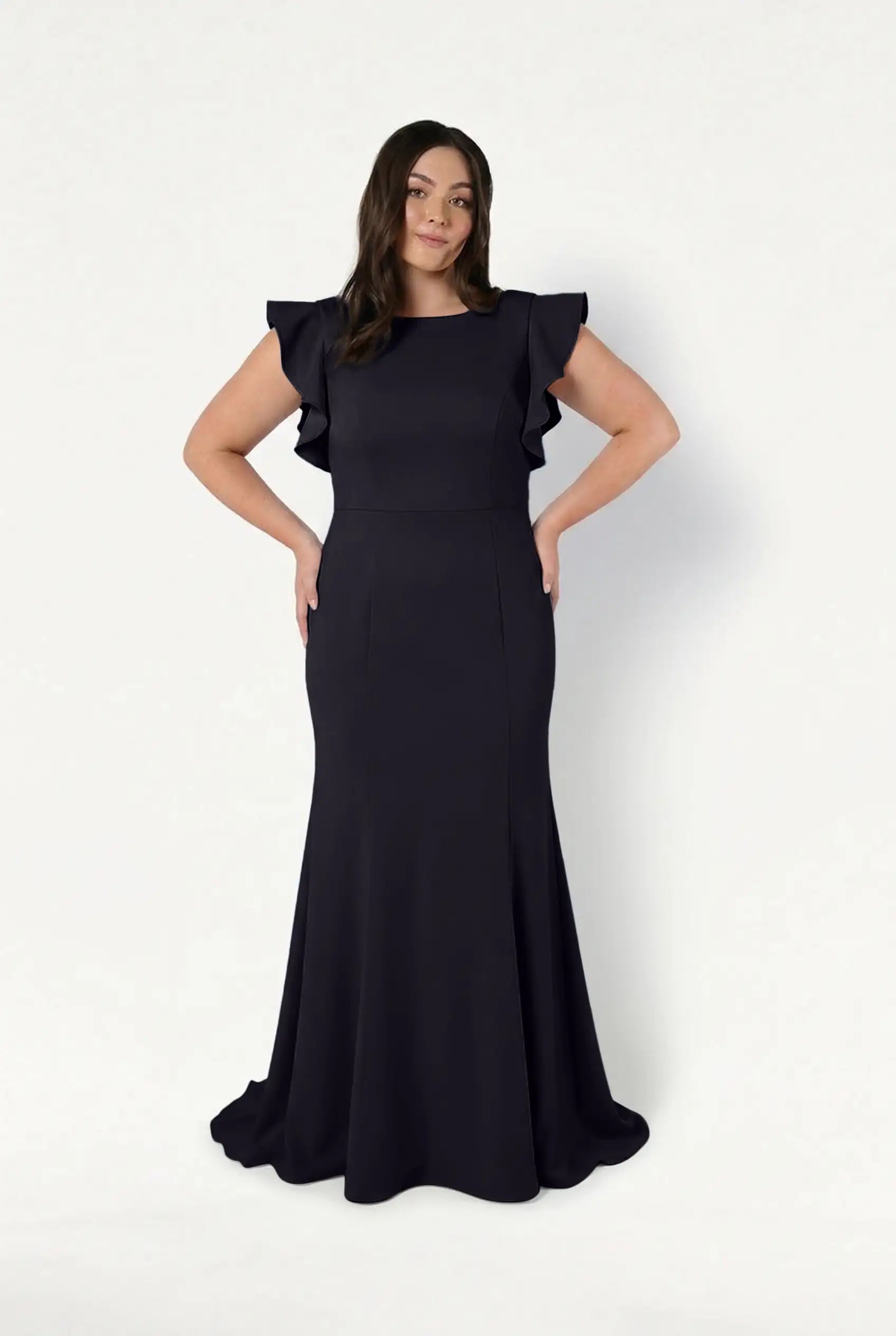 A woman models the TH&TH Cecelia Moss Crepe Bridesmaid Dress in Black—a floor-length, fitted gown with ruffled short sleeves—standing against a white background with hands on hips and a soft smile.