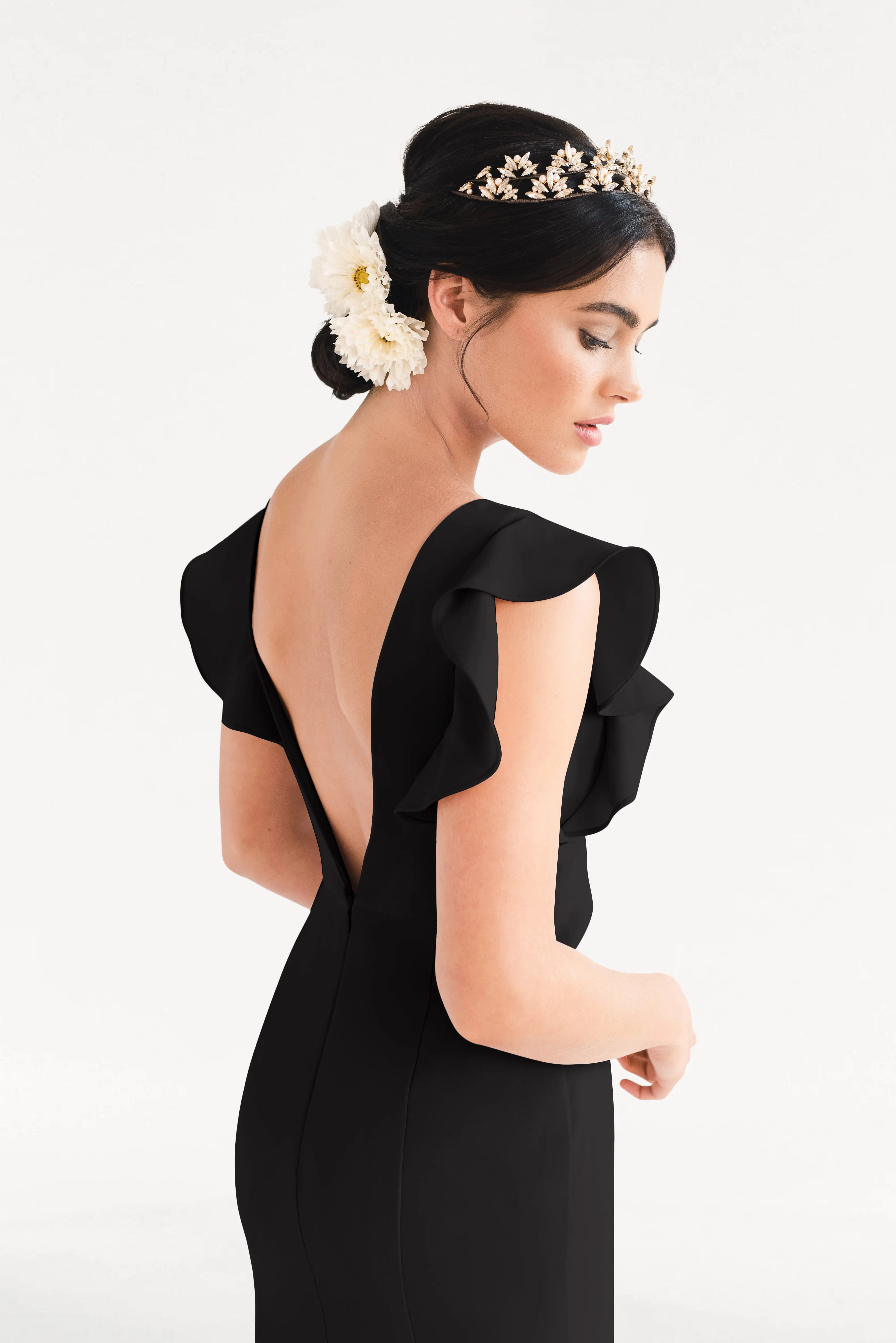 A woman in the TH&TH Cecelia Moss Crepe Bridesmaid Dress in Black stands turned to the side, showcasing its modern backless design and ruffled sleeves. She wears a floral hairpiece, a gold tiara, and her dark hair is styled in a low bun against a white background.
