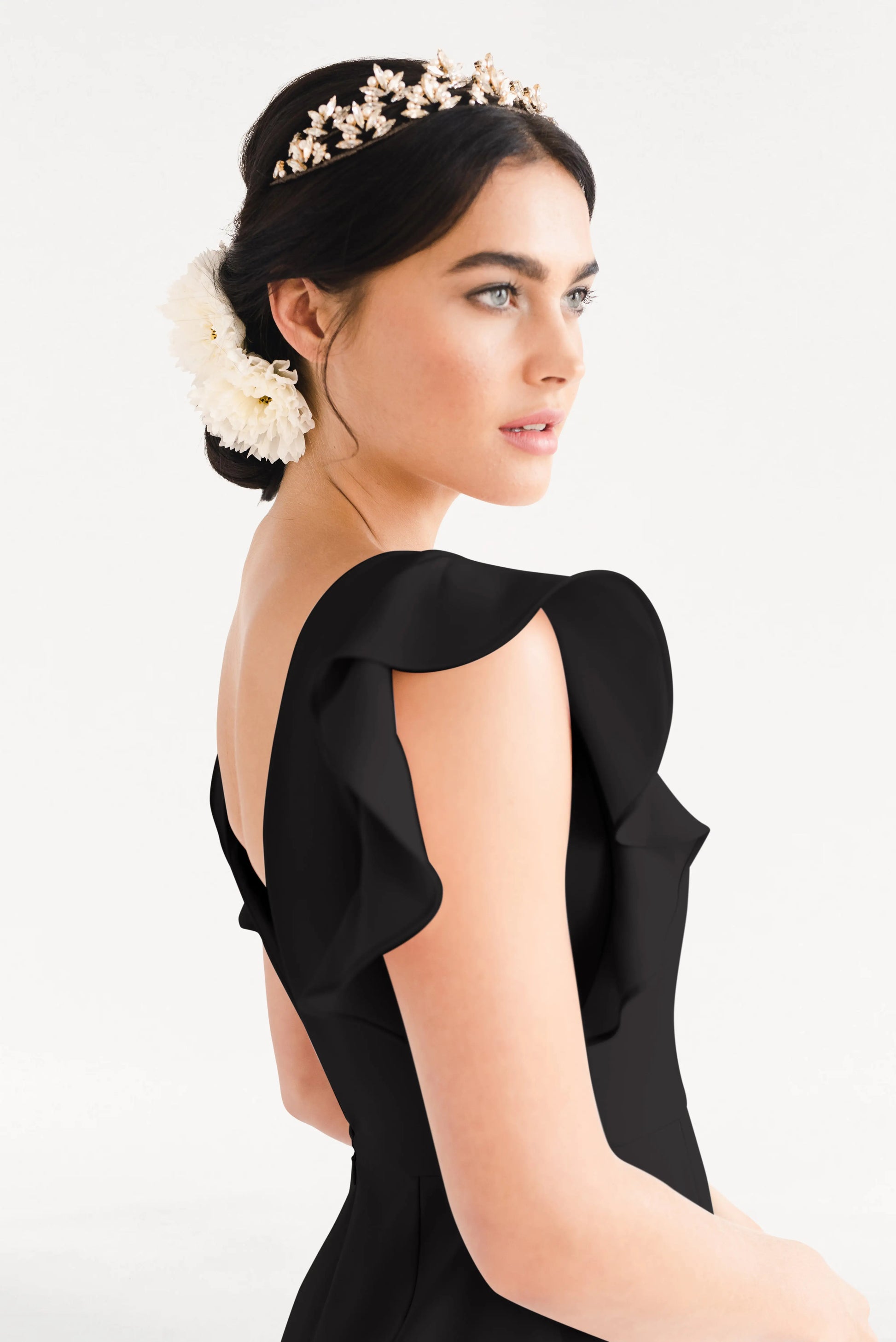A woman with fair skin and dark hair wears the TH&TH Cecelia Moss Crepe Bridesmaid Dress in Black, a gold floral headpiece, and a white flower in her hair, looking to the side against a plain white background.
