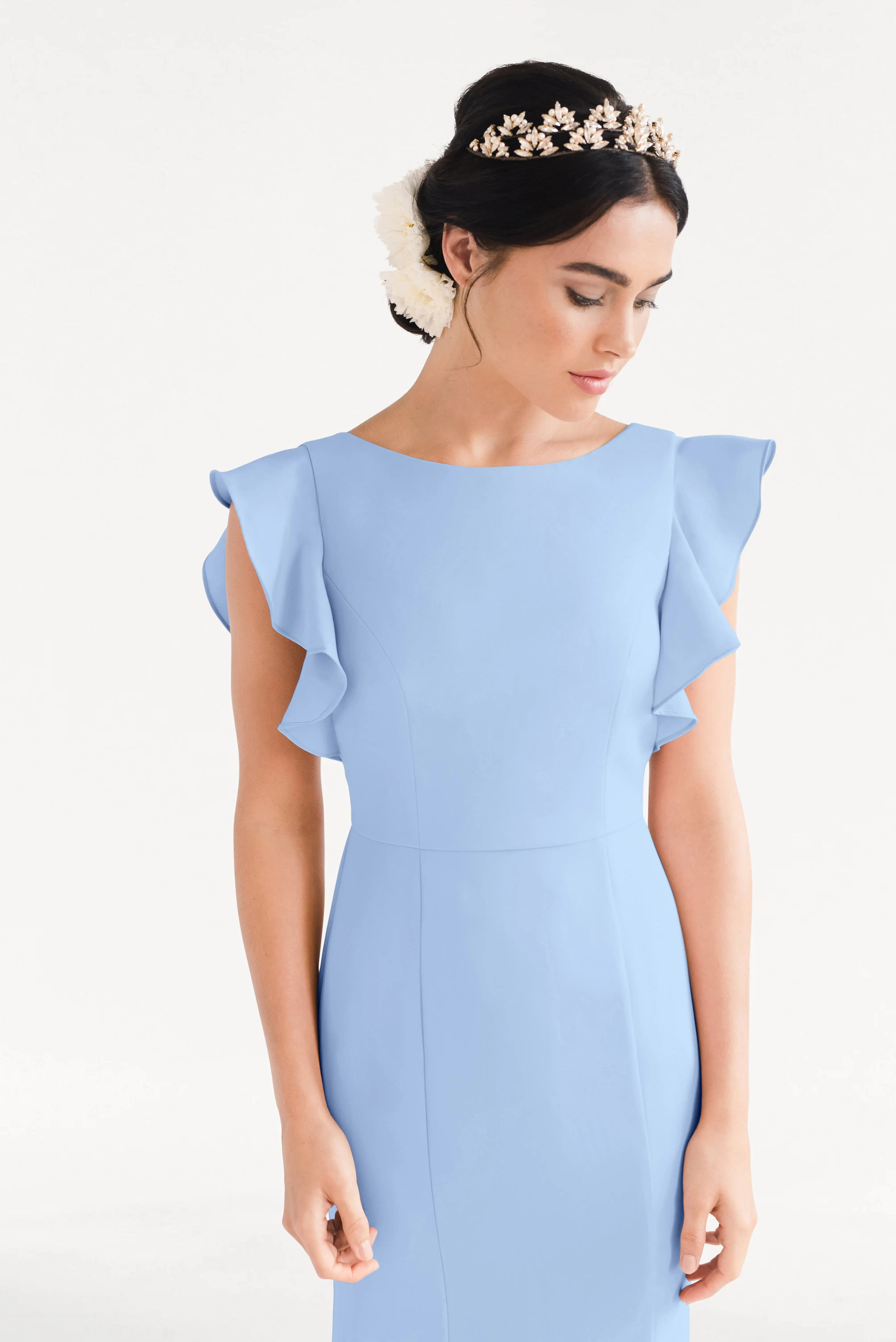 A woman in the TH&TH Cecelia Moss Crepe Bridesmaid Dress in Dusty Blue, sized inclusively, stands against a white background. The gown features ruffled sleeves, and her dark updo is adorned with a floral headpiece.