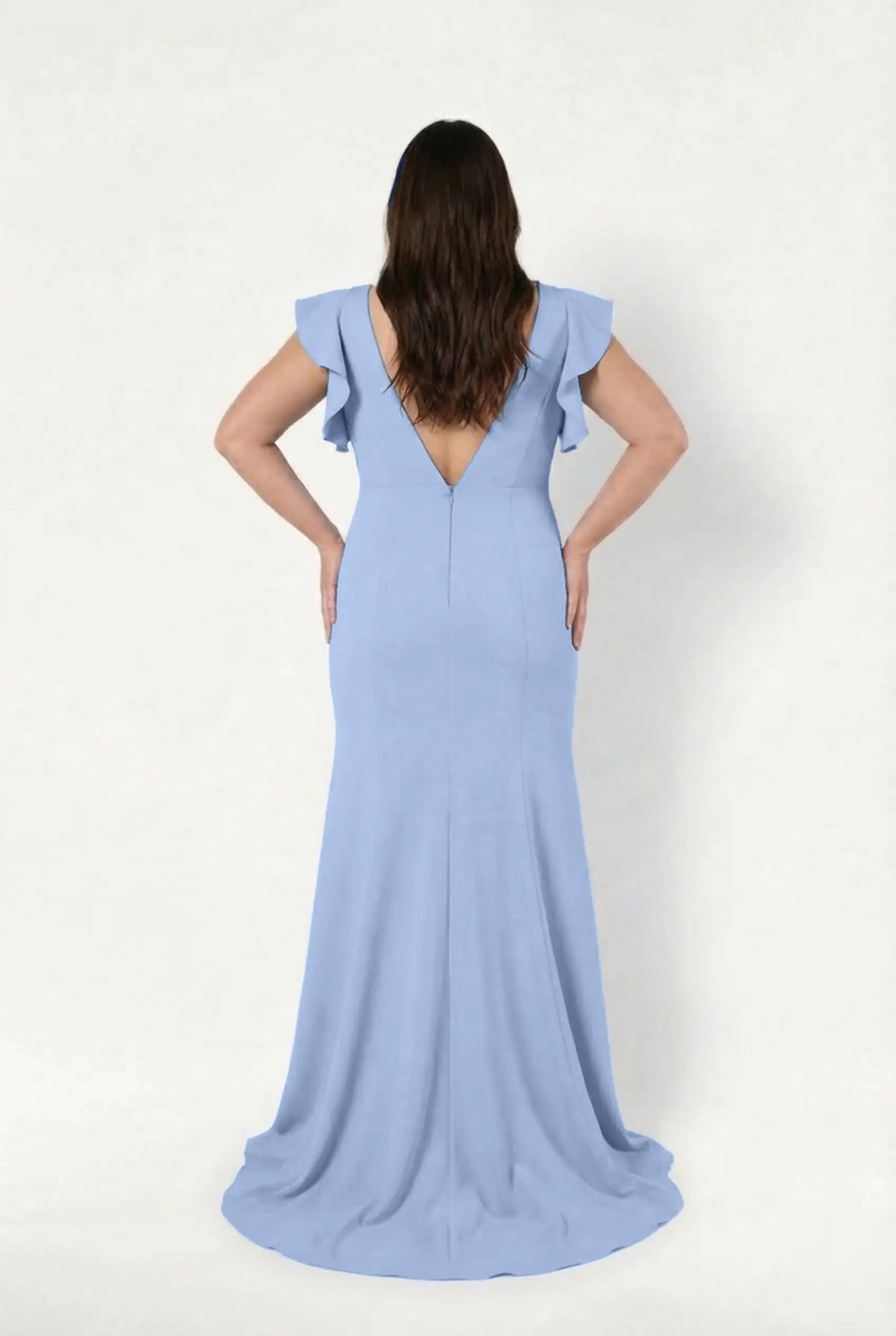 A woman with long brown hair faces away, wearing the TH&TH Cecelia Moss Crepe Bridesmaid Dress in Dusty Blue, featuring a deep V-shaped back and ruffled short sleeves, set against a plain white background.