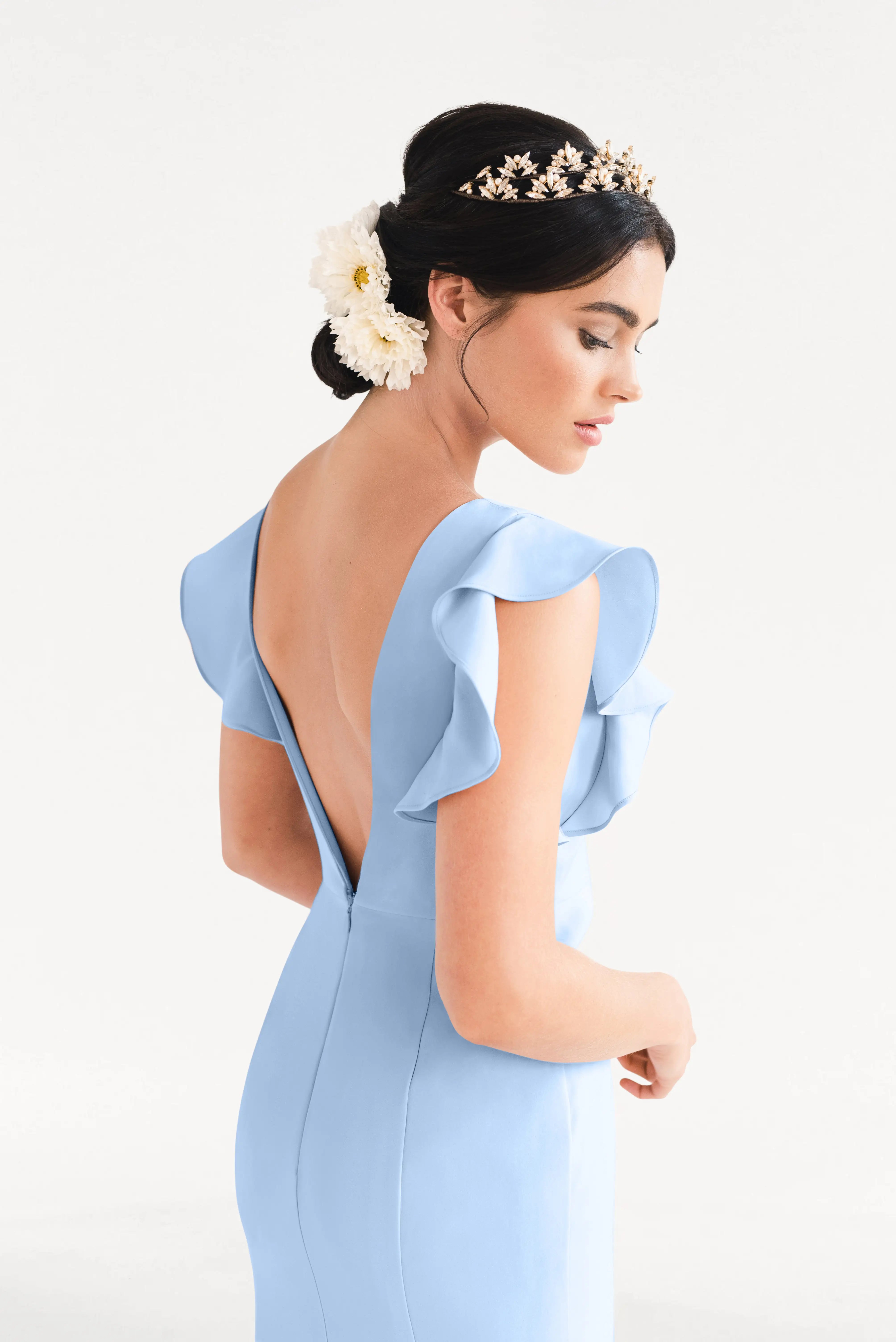 A woman shown from behind on a white background wears the TH&TH Cecelia Moss Crepe Bridesmaid Dress in Dusty Blue, featuring ruffle sleeves, a gold crystal tiara, and white flowers in her dark updo.