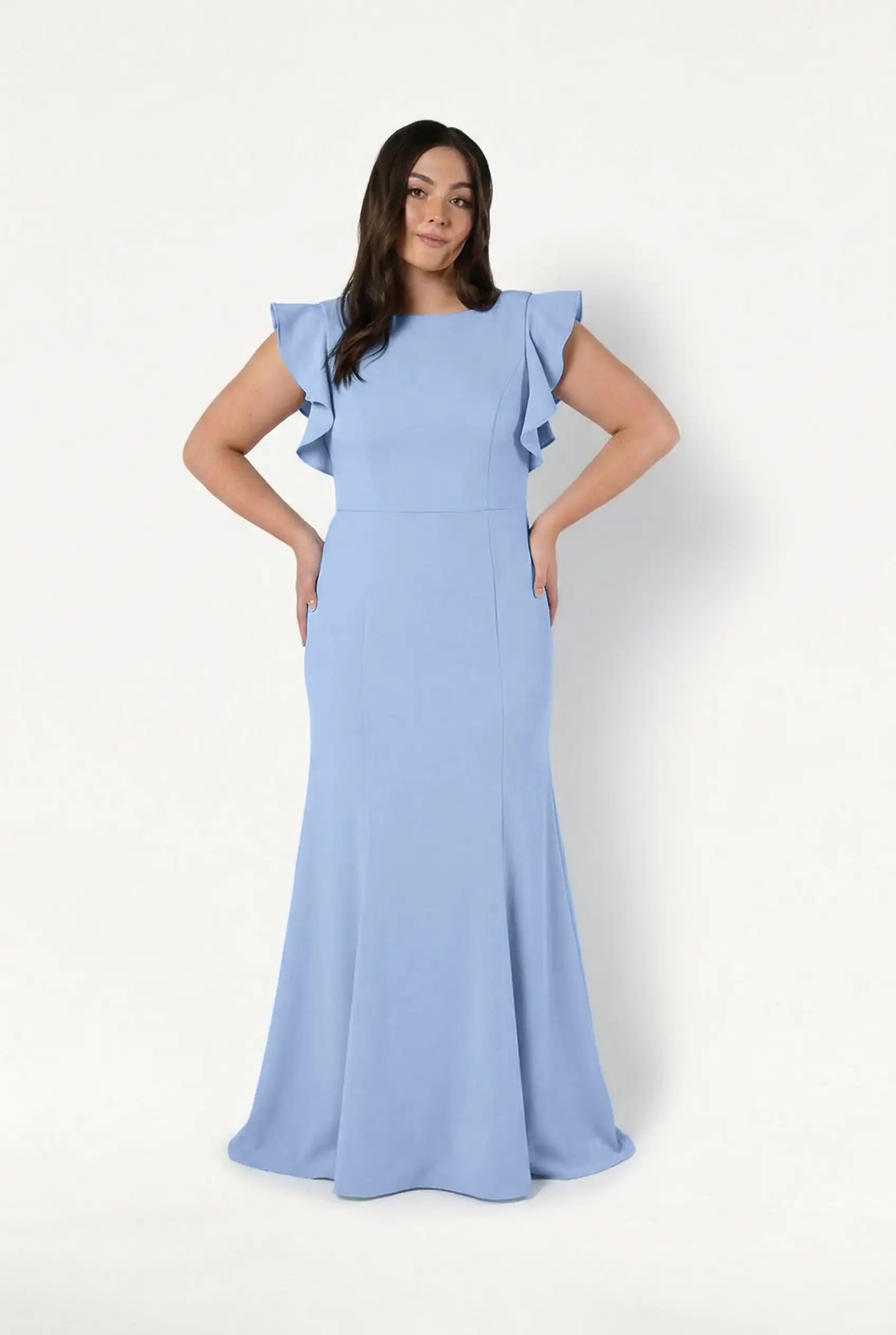A woman with long dark hair and a confident look models the TH&TH Cecelia Moss Crepe Bridesmaid Dress in Dusty Blue—a size-inclusive, floor-length gown with ruffled short sleeves—against a white background.