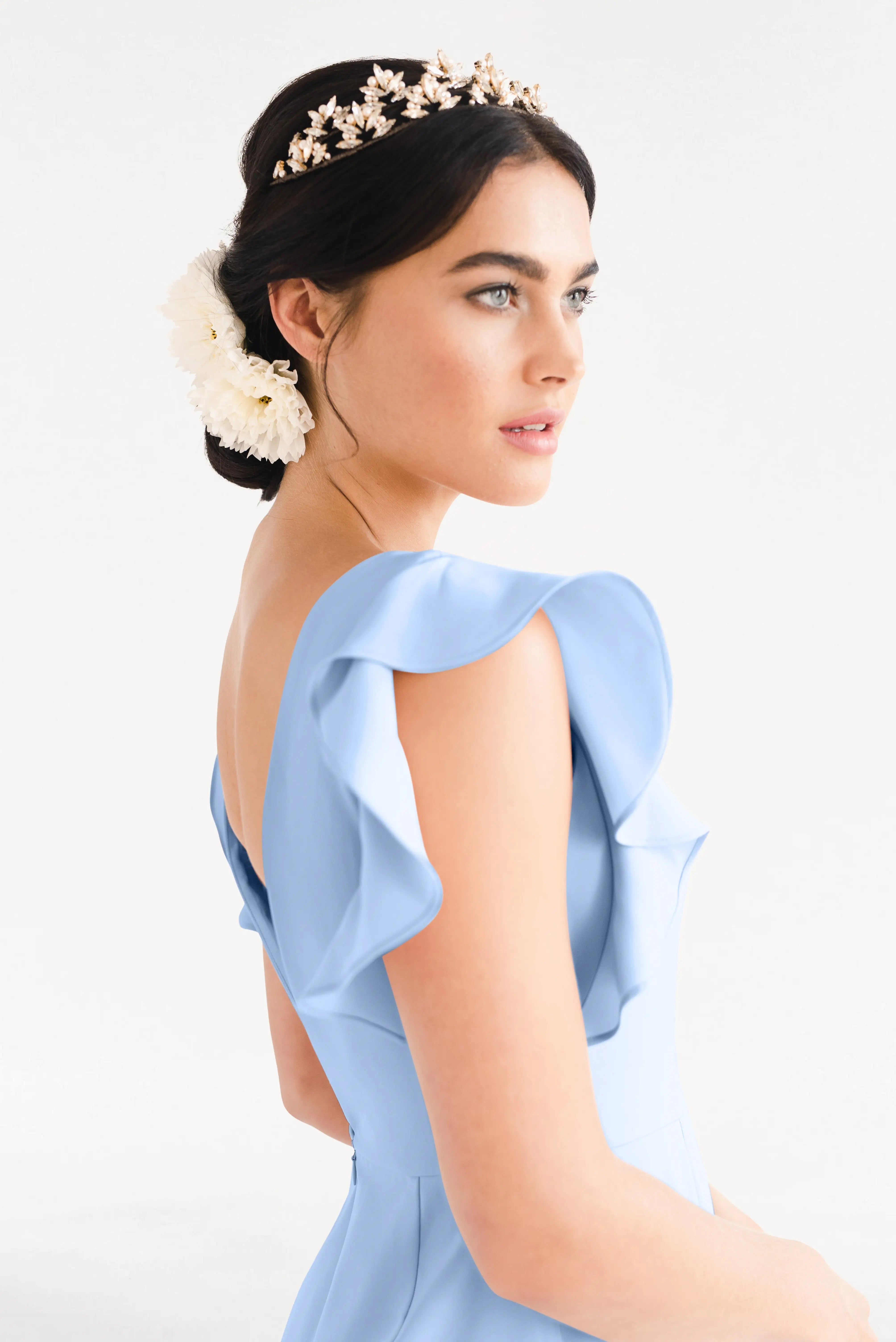 Woman in the TH&TH Cecelia Moss Crepe Bridesmaid Dress in Dusty Blue, a size-inclusive gown with ruffled sleeves, styled with a gold floral headpiece and white flower in her updo, poses elegantly against a plain light background.