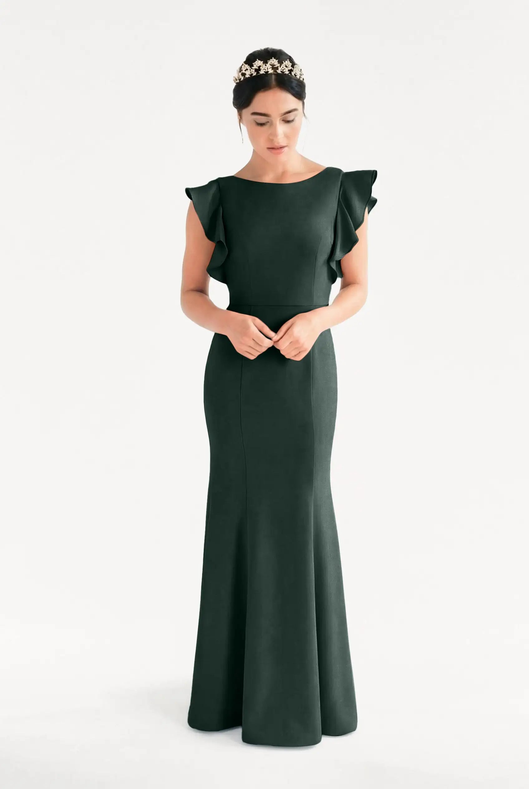 A woman stands against a plain white background wearing TH&TH’s Cecelia Moss Crepe Bridesmaid Dress in Forest Green, featuring ruffled shoulders. She looks down with hands clasped and wears a jeweled headband.