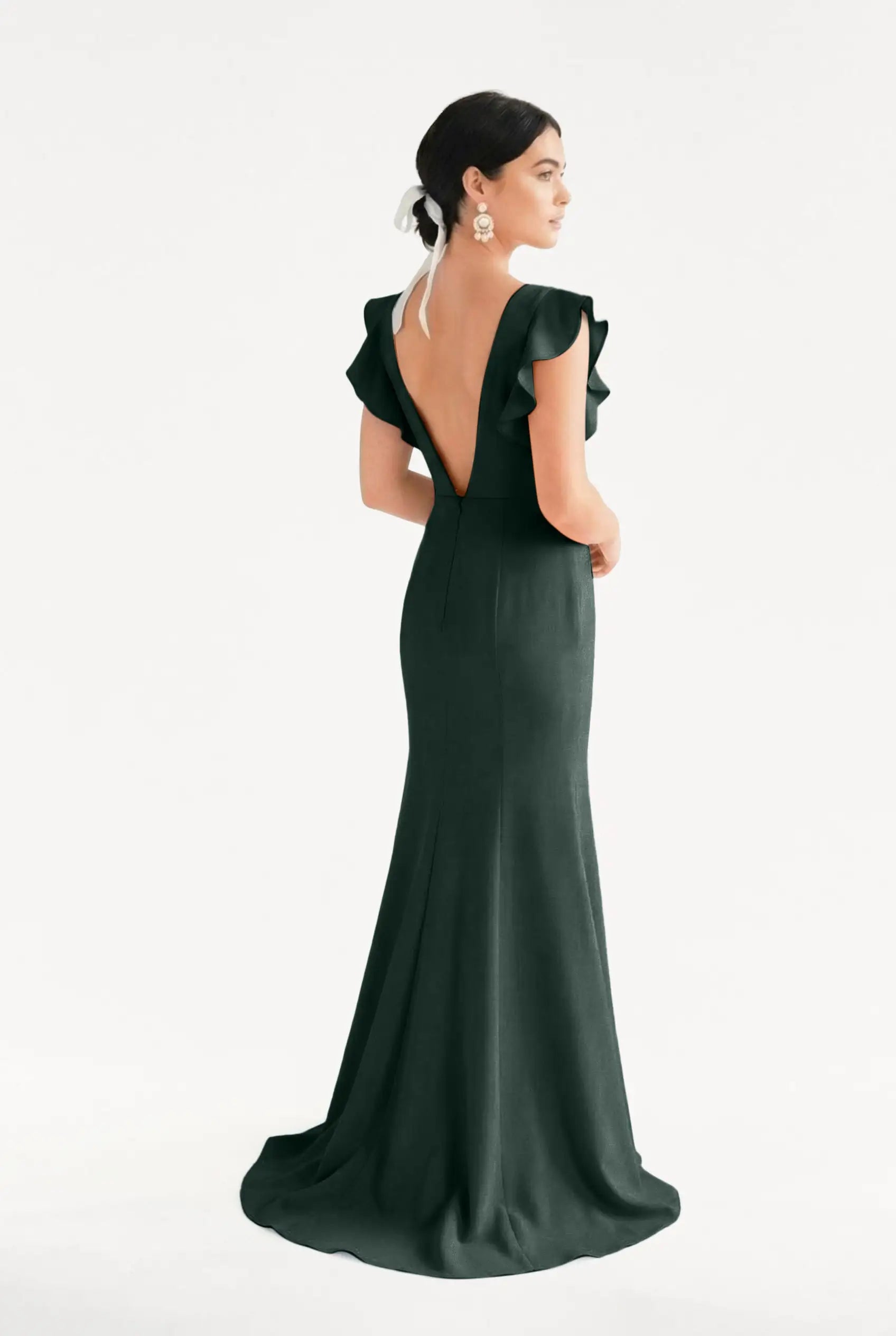 A woman faces away in the TH&TH Cecelia Moss Crepe Bridesmaid Dress in Forest Green—a floor-length, size-inclusive gown with ruffled sleeves and a deep V-back. Her hair is up with a white ribbon and statement earrings, set against a plain background.
