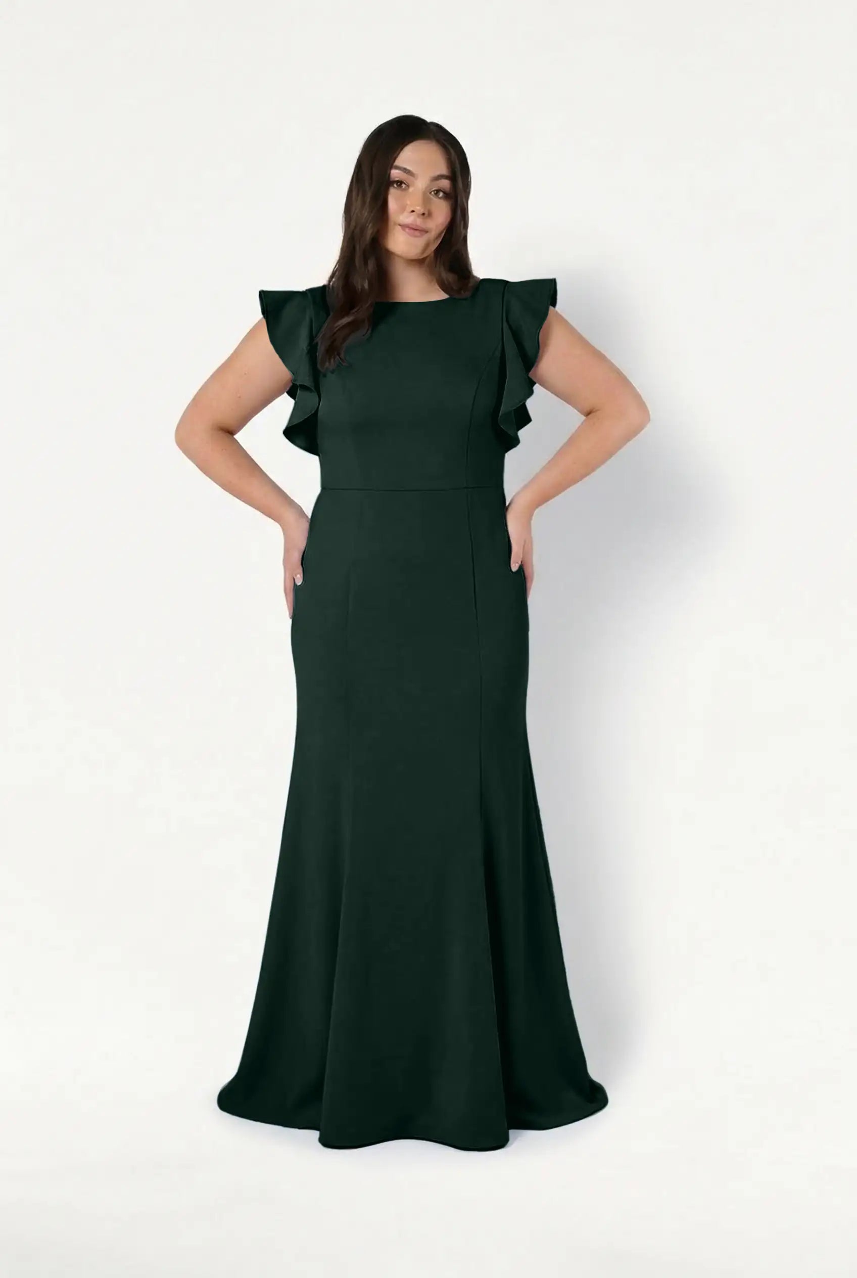 A woman smiles softly with her hands on her hips, wearing the floor-length TH&TH Cecelia Moss Crepe Bridesmaid Dress in Forest Green, featuring ruffled short sleeves, against a plain white background.