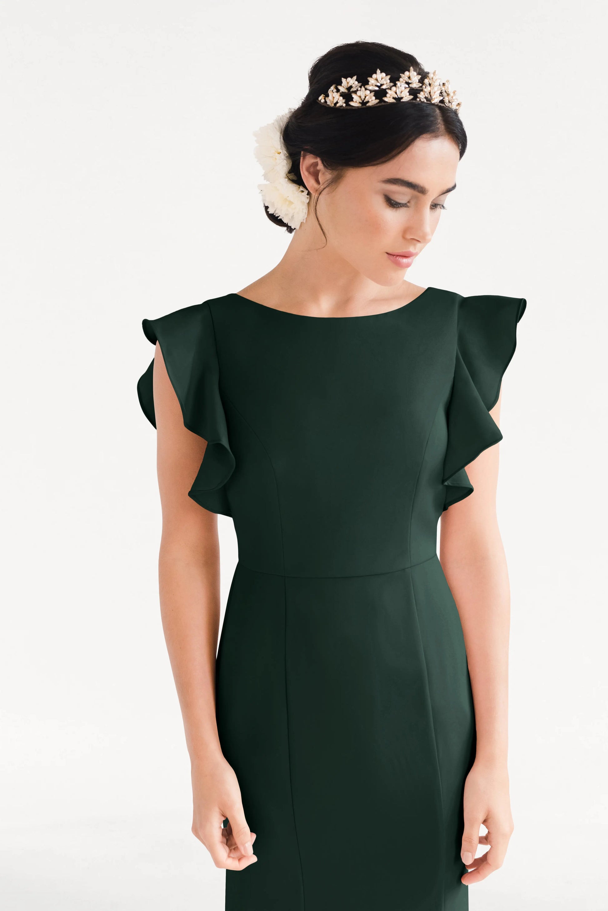 A woman wearing the TH&TH Cecelia Moss Crepe Bridesmaid Dress in Forest Green with ruffled sleeves and a floral headpiece looks down against a plain white background.