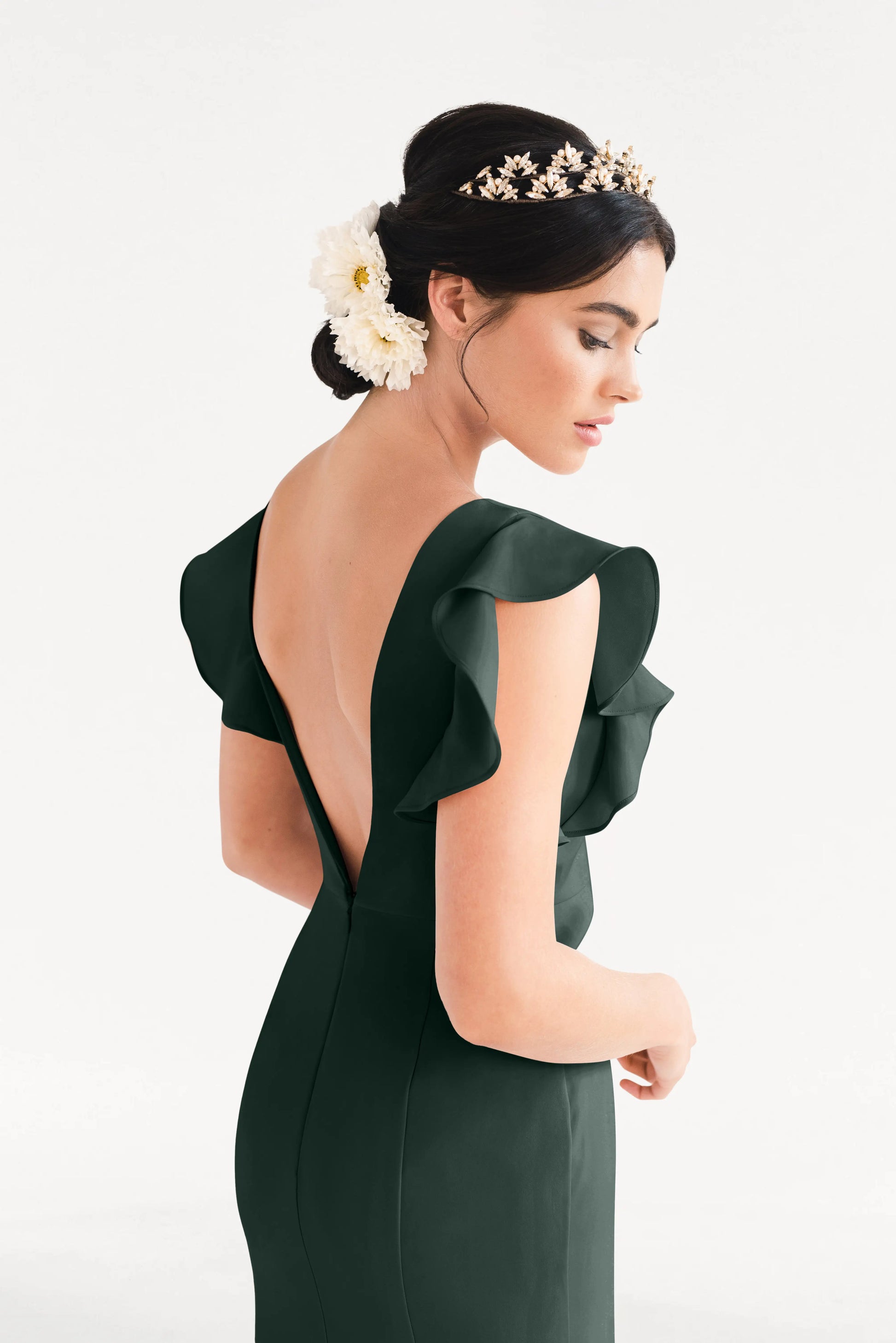A woman in the Cecelia Moss Crepe Bridesmaid Dress in Forest Green by TH&TH stands in profile, featuring ruffled sleeves and an open back. She wears a gold floral headpiece with flowers in her hair against a plain white background.