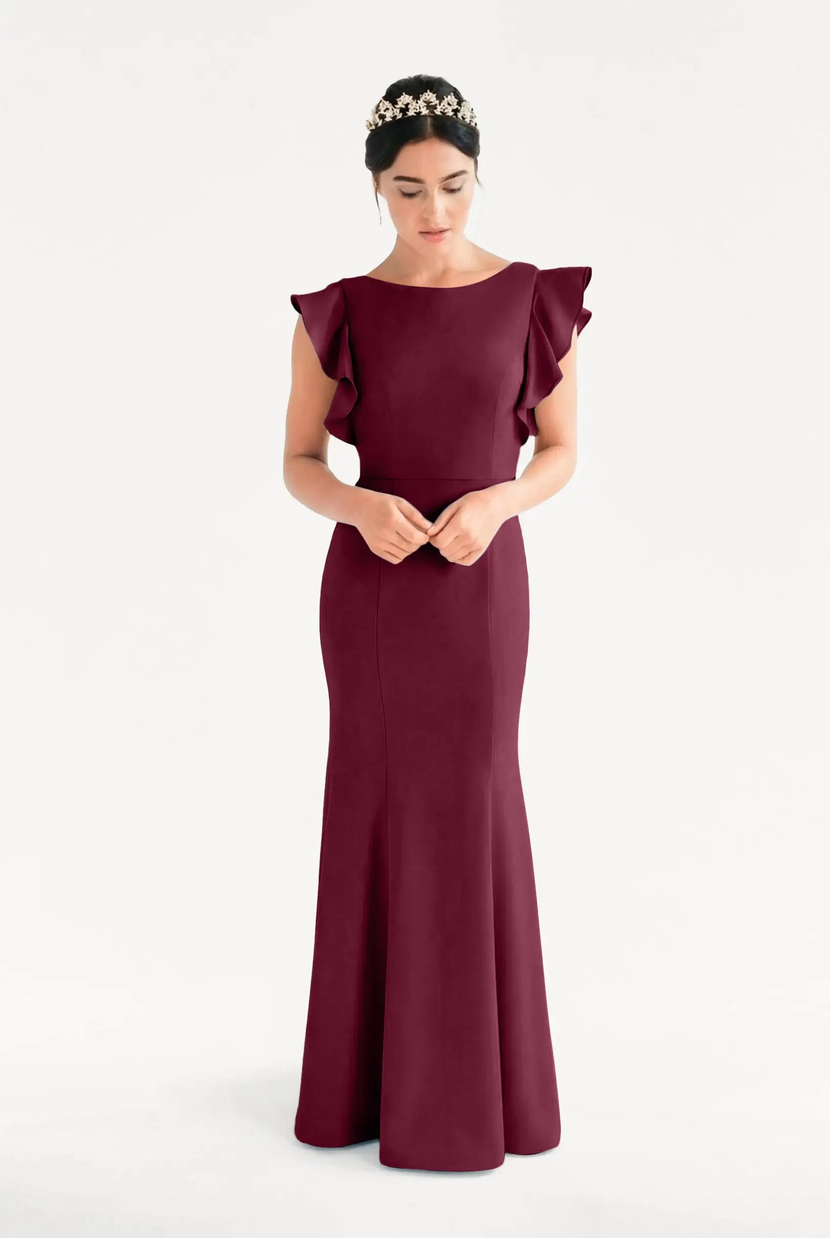 A woman wears the Cecelia Moss Crepe Bridesmaid Dress in Merlot by TH&TH—a floor-length, sleeveless gown with ruffled shoulders—standing against a white background, looking down with hands clasped and a jeweled headband in her hair.