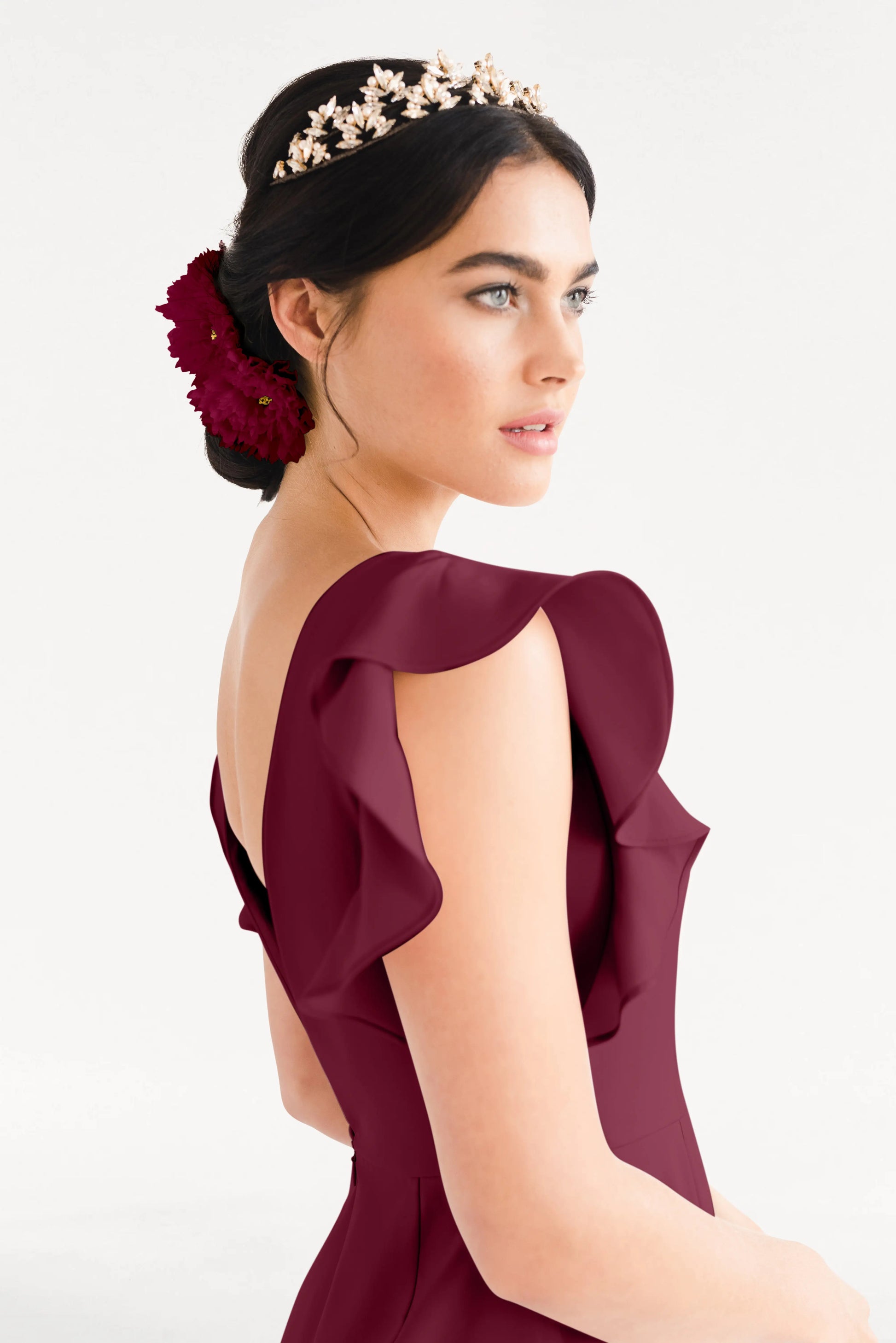 A woman with dark hair in a low bun adorned with red flowers and a gold headpiece wears the TH&TH Cecelia Moss Crepe Bridesmaid Dress in Merlot, featuring ruffled shoulders, as she looks to the side against a plain background.