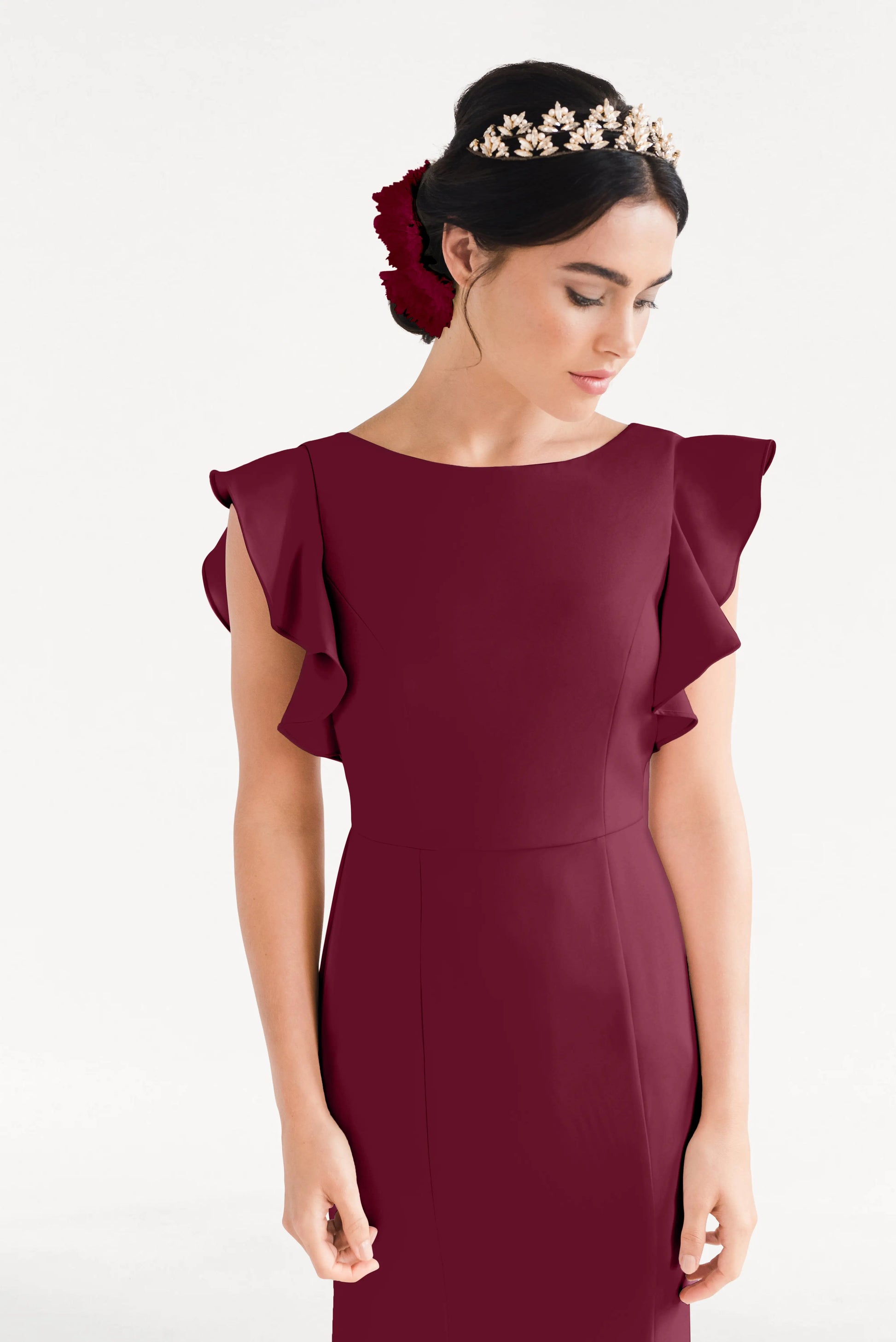 A woman wears the TH&TH Cecelia Moss Crepe Bridesmaid Dress in Merlot, featuring ruffled sleeves. Her hair is styled in an elegant updo with red flowers and a floral headpiece. She looks downward against a plain white background.