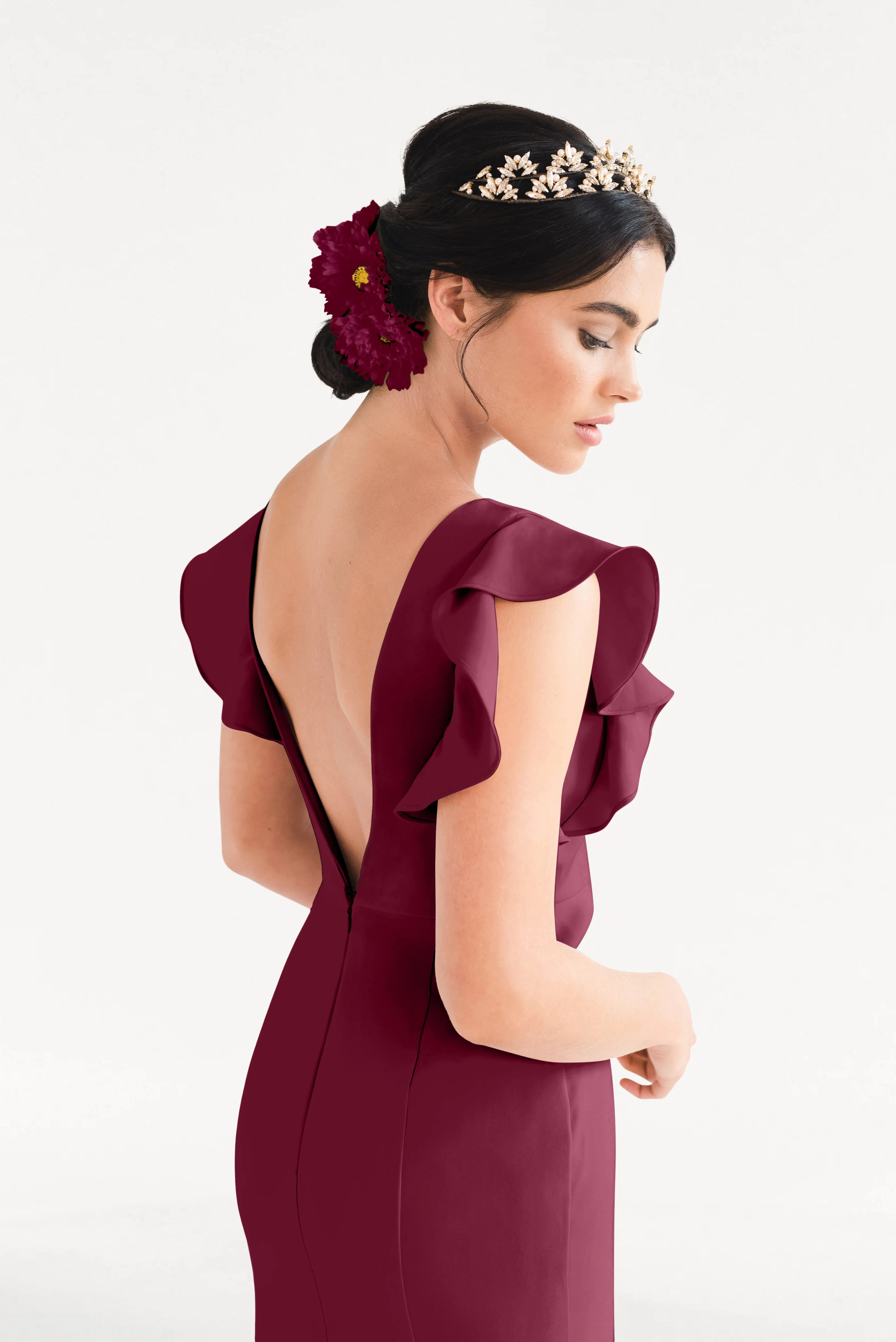 A woman wears the TH&TH Cecelia Moss Crepe Bridesmaid Dress in Merlot, featuring ruffled sleeves and a backless design, styled with a golden floral headpiece and large red flower, against a plain white background.