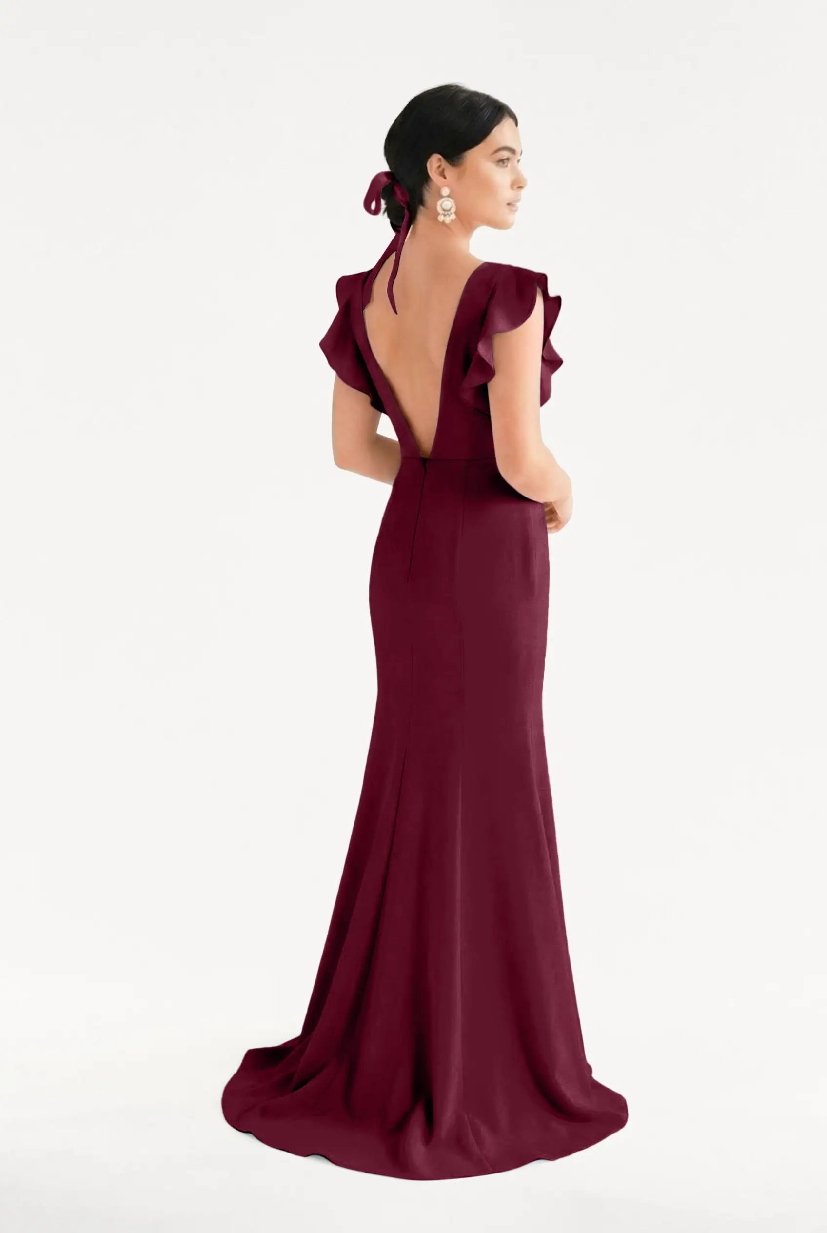 A woman models the TH&TH Cecelia Moss Crepe Bridesmaid Dress in Merlot, a size-inclusive, floor-length gown with ruffled sleeves and a low V-back. She wears her dark hair up with a matching ribbon and statement earrings against a white background.