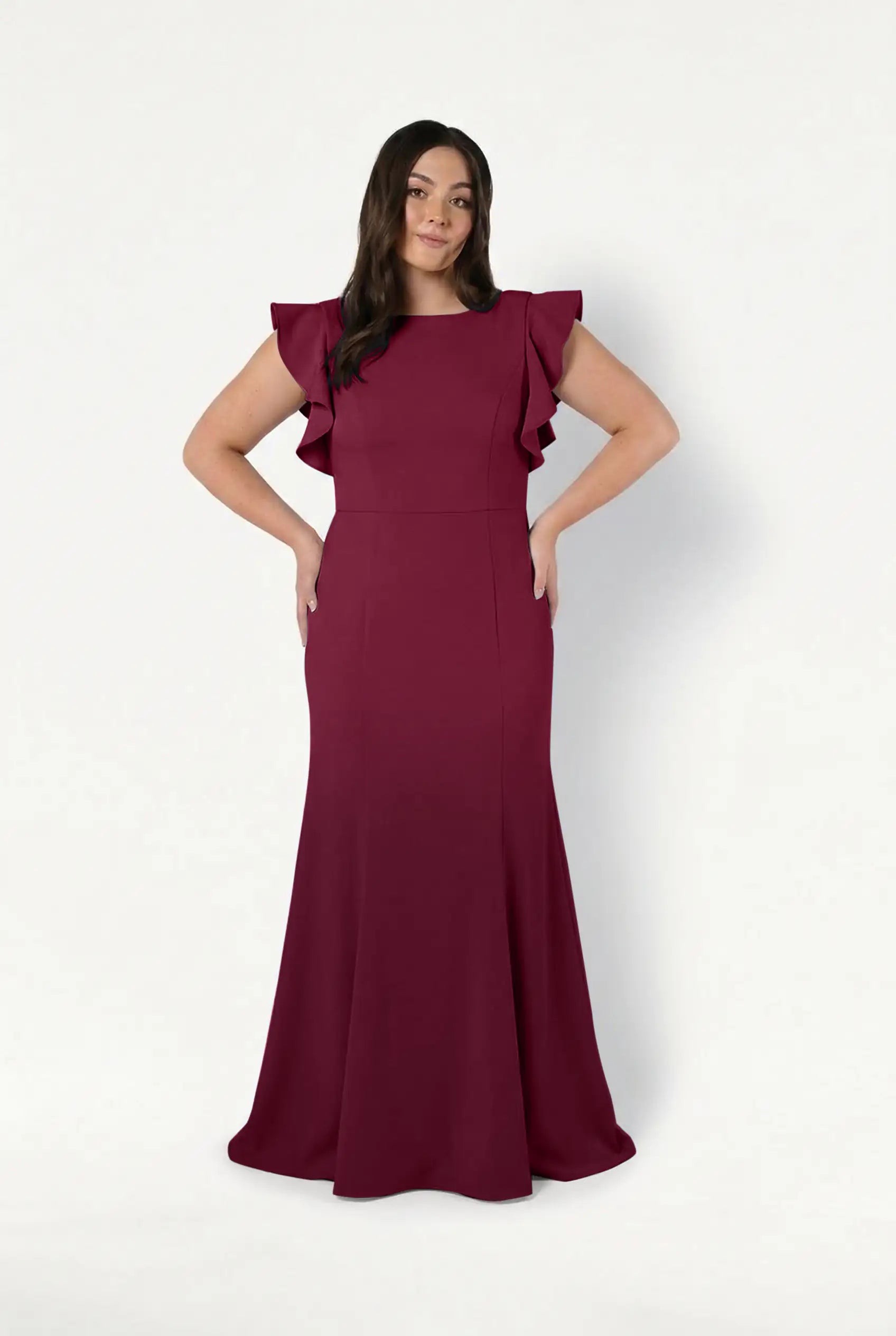A woman smiles with her hands on her hips, modeling the TH&TH Cecelia Moss Crepe Bridesmaid Dress in Merlot—a size-inclusive, sleeveless, floor-length gown with ruffled shoulders—against a plain white background.