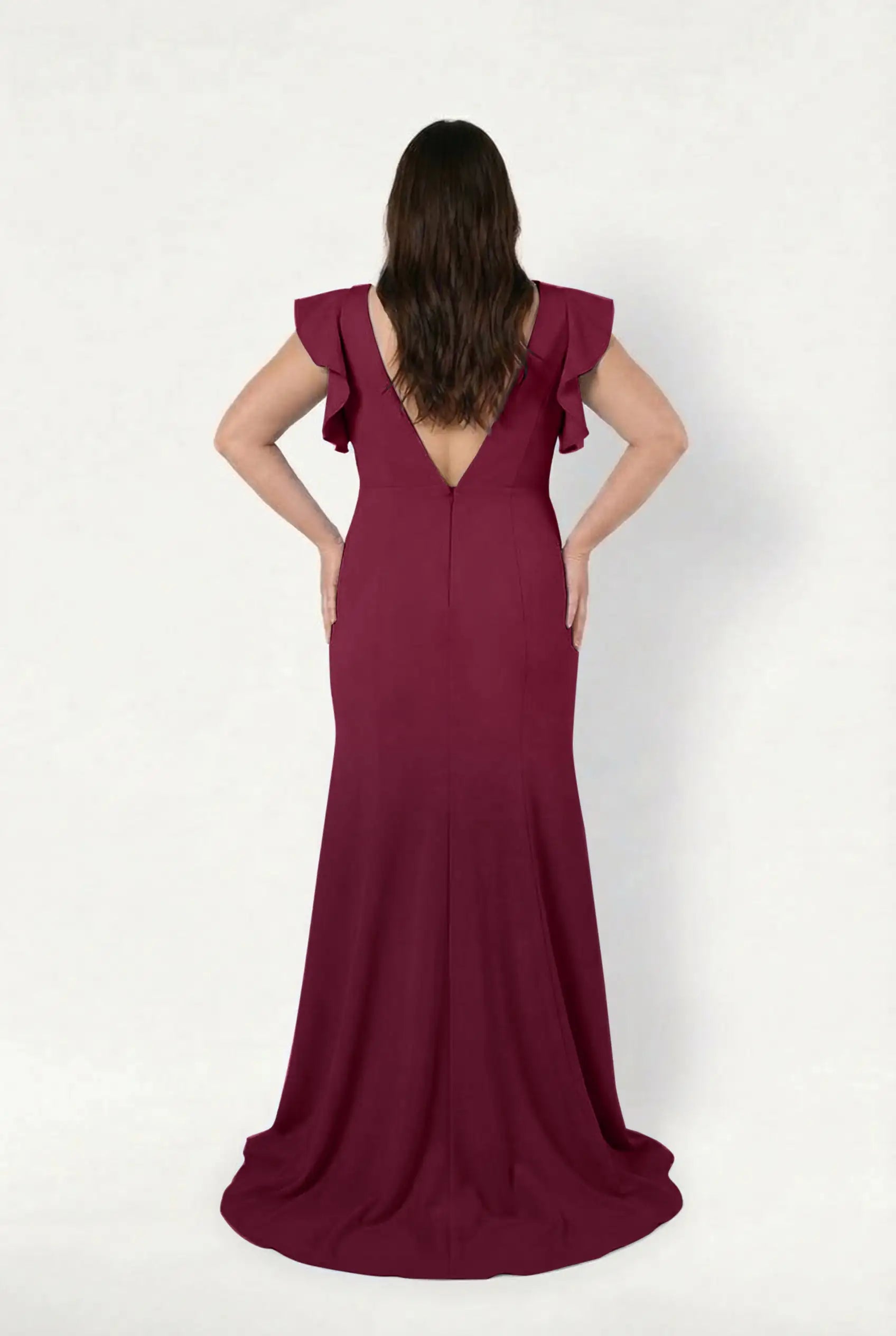 A woman stands facing away, wearing the TH&TH Cecelia Moss Crepe Bridesmaid Dress in Merlot—a floor-length gown with a V-back and ruffled cap sleeves. Her hands are on her hips, long dark hair falling against a plain white backdrop.
