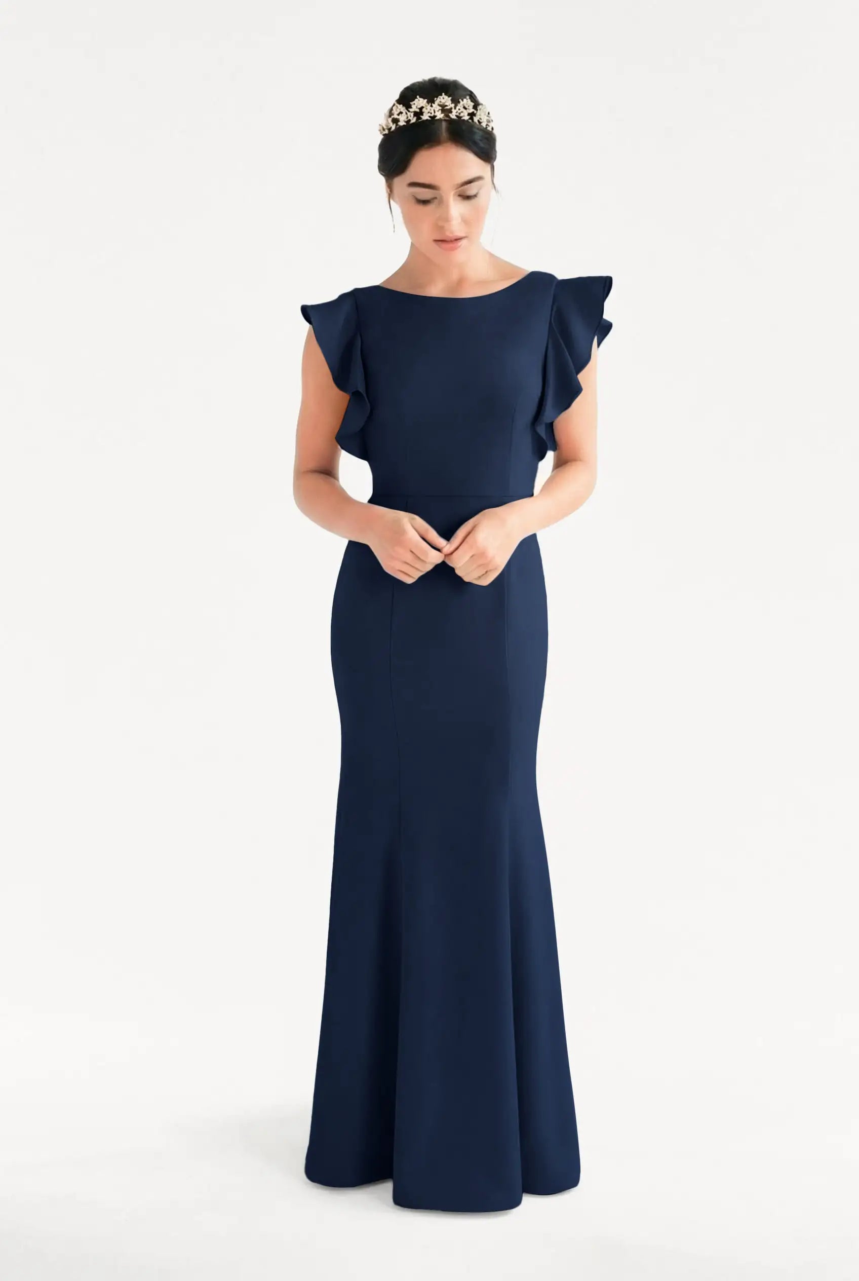 A woman wearing the TH&TH Cecelia Moss Crepe Bridesmaid Dress in Navy—a floor-length, sleeveless gown with ruffled shoulders and a fitted silhouette—stands with hands folded, looking down, accessorized with a floral headband.
