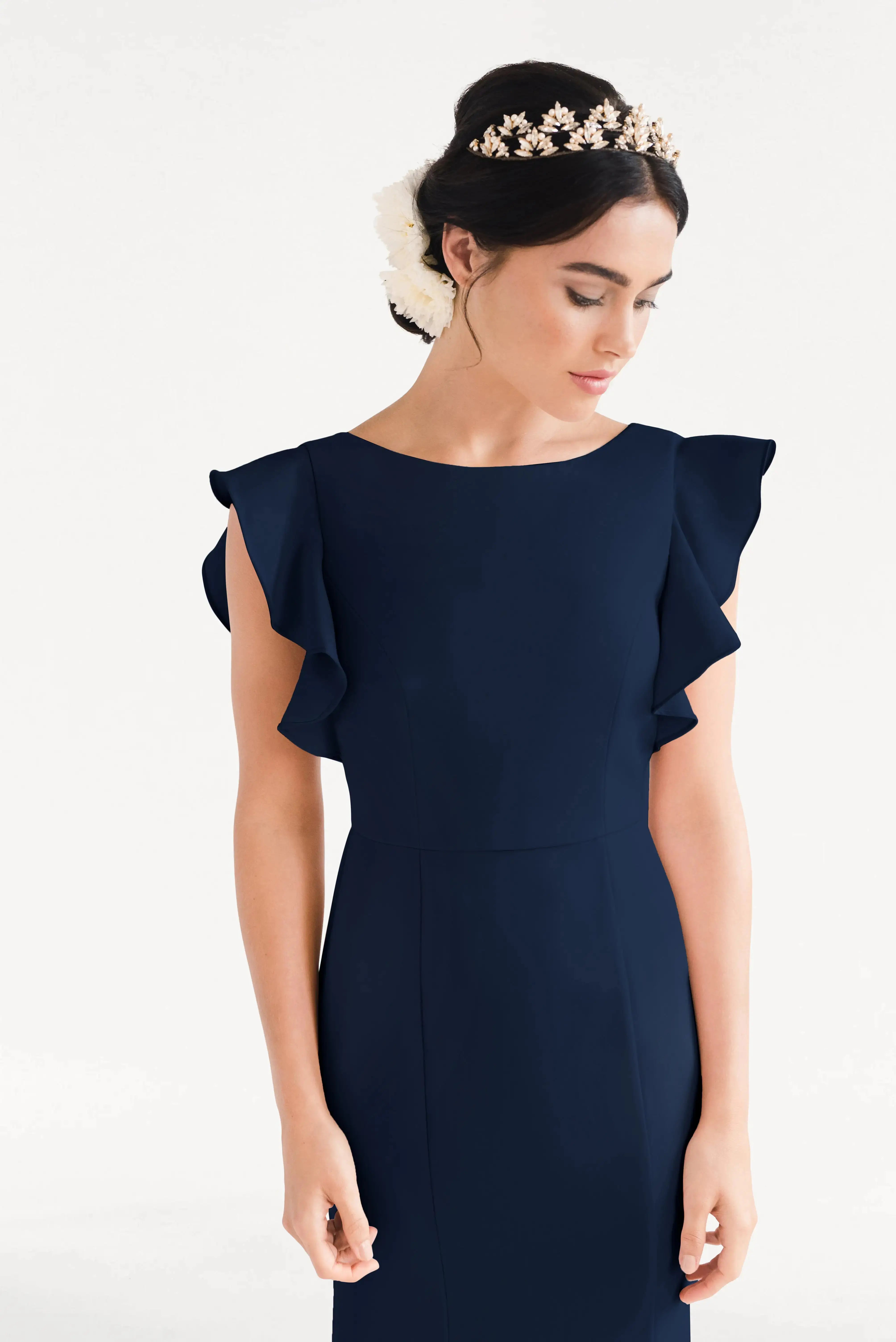 A woman models the TH&TH Cecelia Moss Crepe Bridesmaid Dress in Navy, a size-inclusive gown with ruffled sleeves, paired with a floral headpiece, against a plain white background.