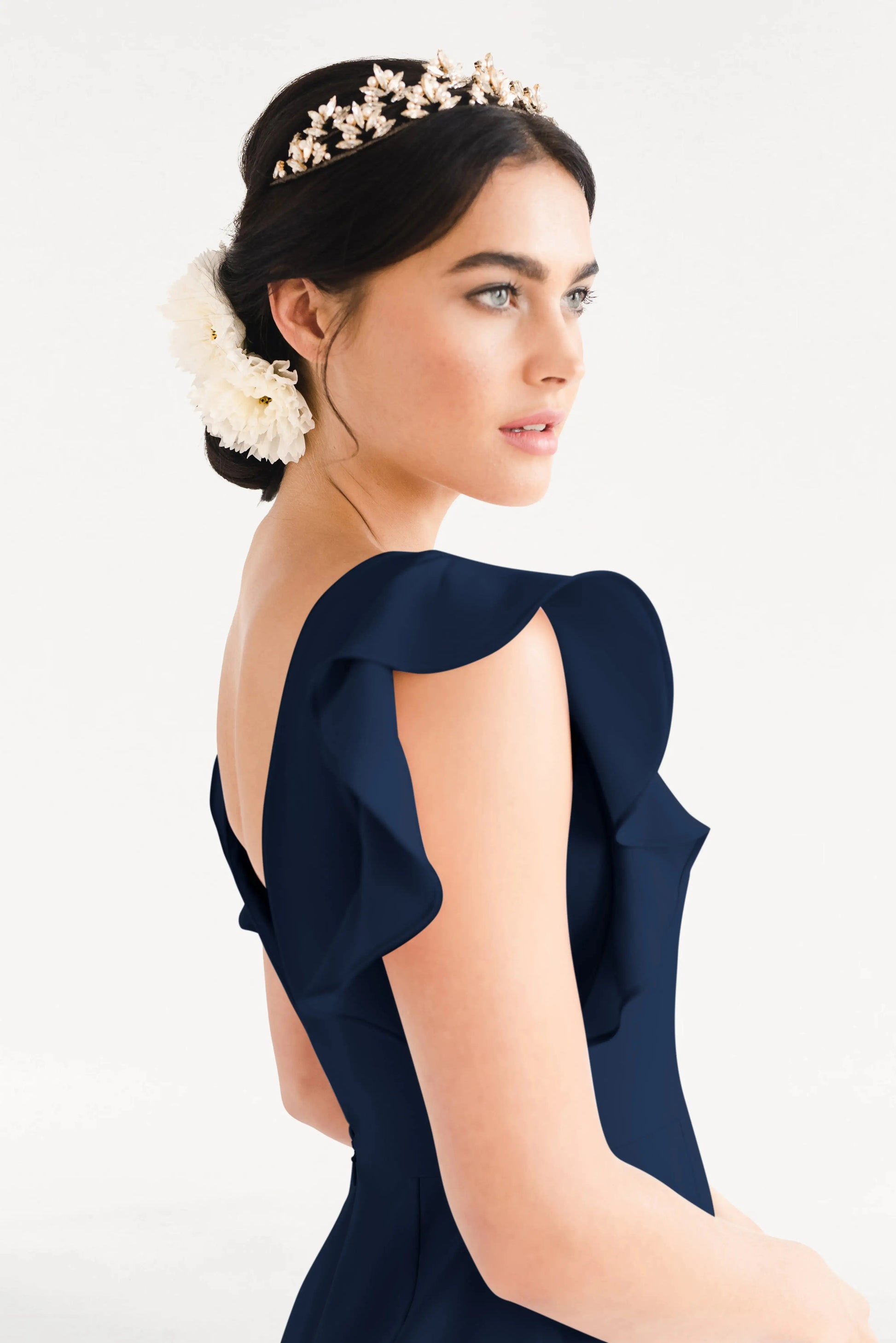 A woman with dark hair in an updo, adorned with a floral headpiece, wears the TH&TH Cecelia Moss Crepe Bridesmaid Dress in Navy with ruffled sleeves. She looks to the side against a plain white background.