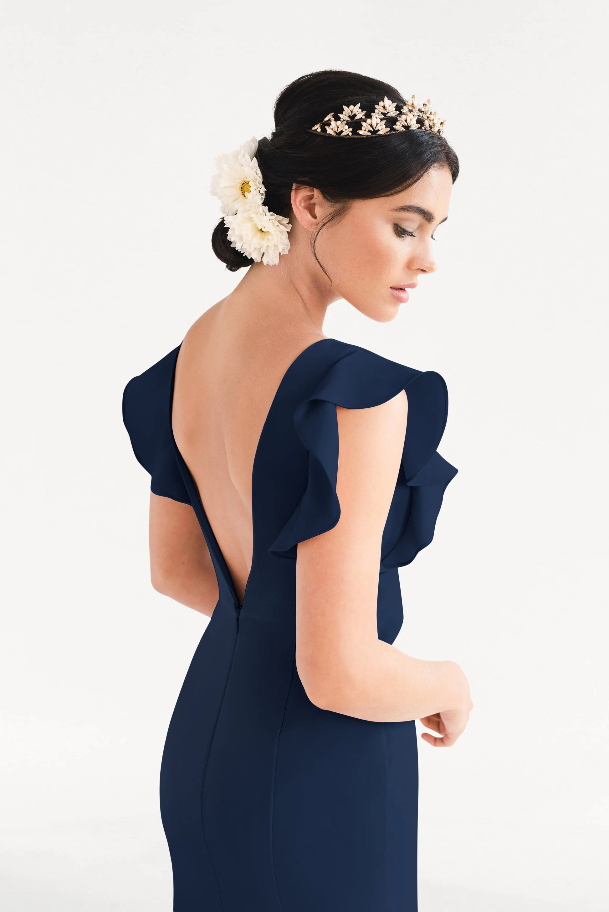 A woman in the TH&TH Cecelia Moss Crepe Bridesmaid Dress in Navy, featuring ruffled sleeves and a deep V-back, with a floral headpiece and large white flowers in her hair, looks down against a plain white background.