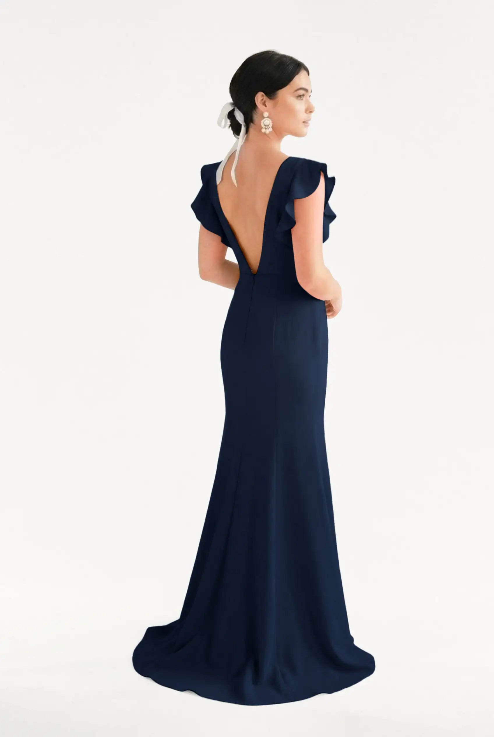 A woman stands against a white background wearing the TH&TH Cecelia Moss Crepe Bridesmaid Dress in Navy, featuring ruffled cap sleeves and a deep V-back. She accessorizes with earrings, a white hair piece, and has her dark hair styled up.