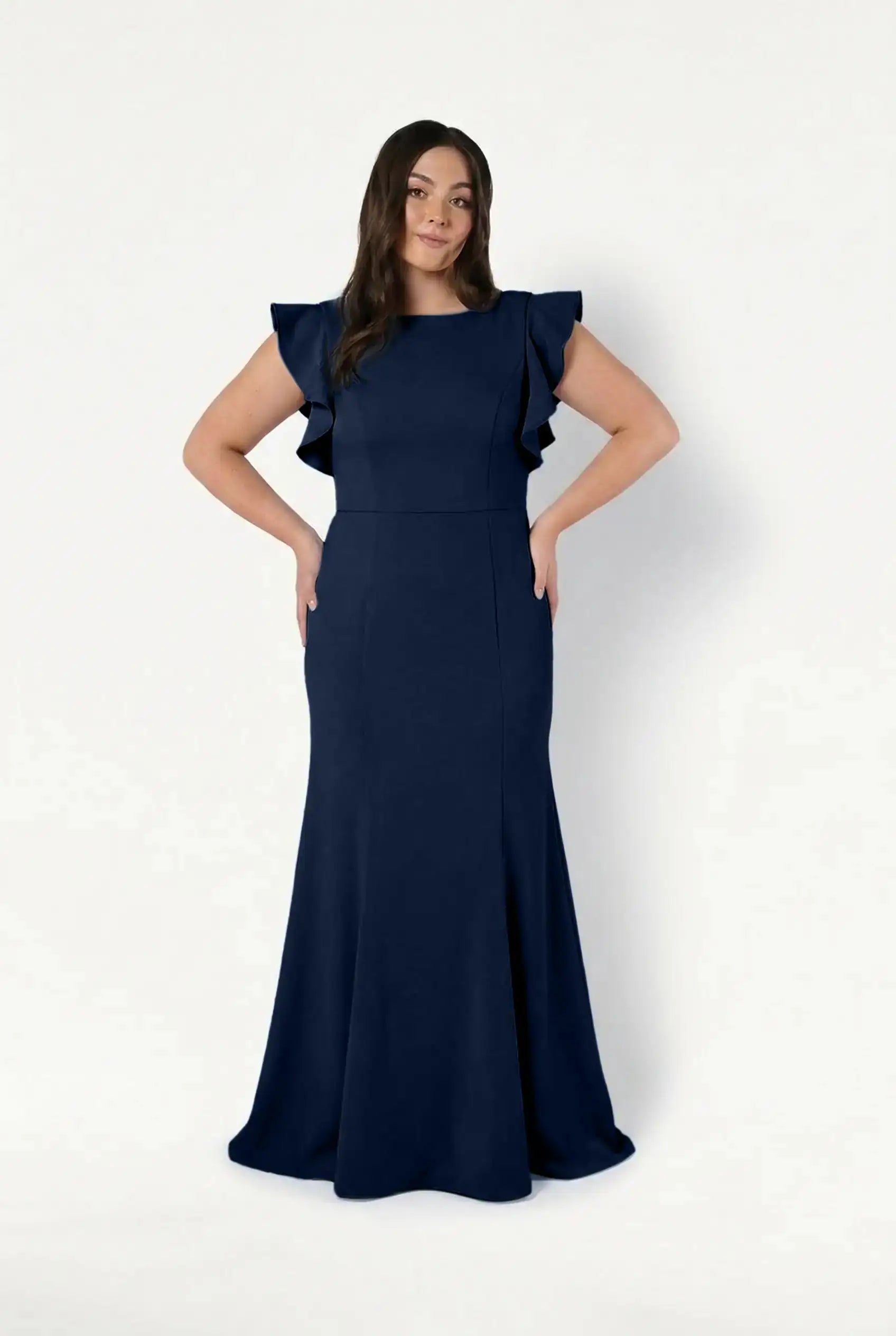 A woman with long, wavy dark hair stands against a white background wearing the TH&TH Cecelia Moss Crepe Bridesmaid Dress in Navy. The floor-length dress features ruffled short sleeves, and she smiles slightly with her hands on her hips.