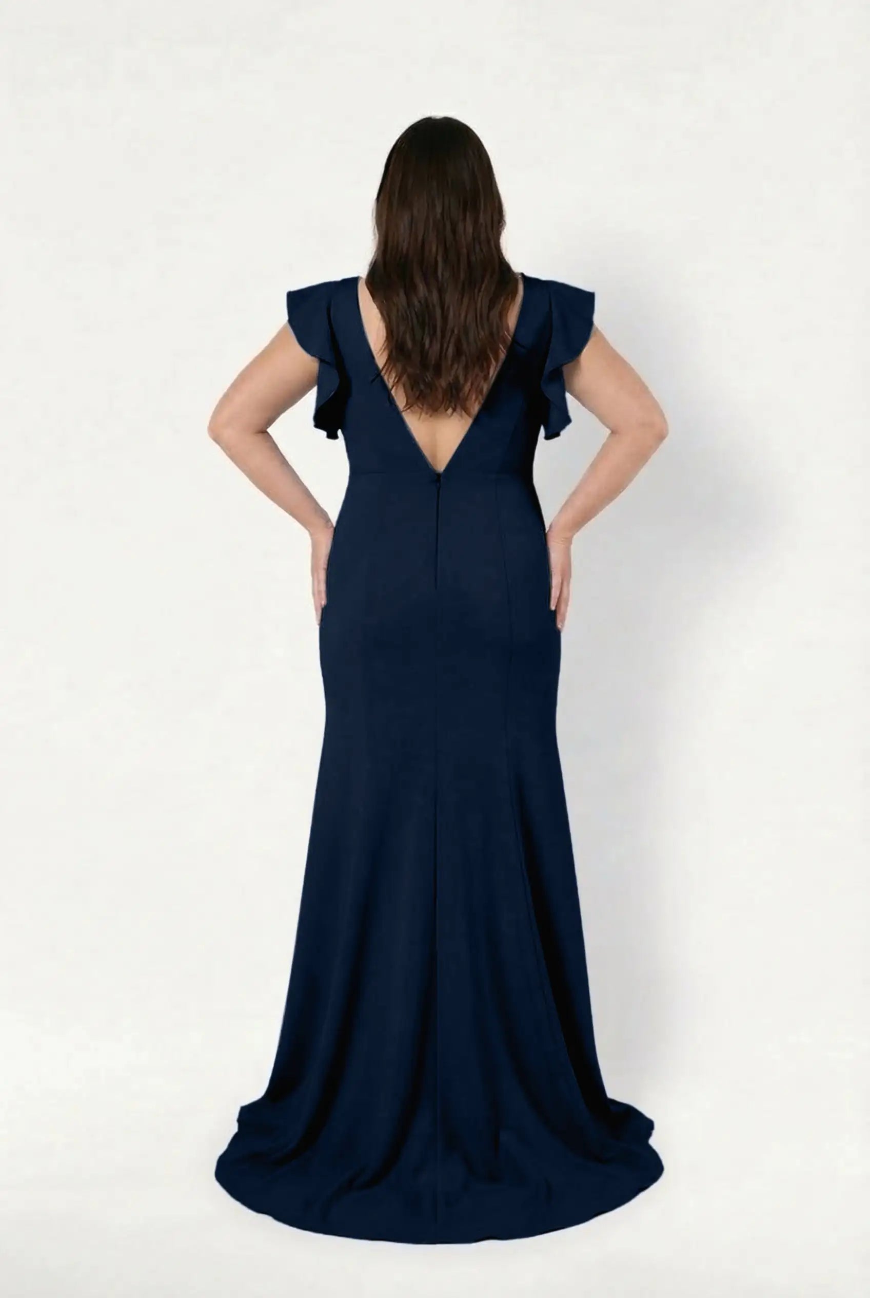 A woman stands with hands on hips, facing away, wearing the Cecelia Moss Crepe Bridesmaid Dress in Navy by TH&TH. The floor-length gown has ruffled sleeves, a deep V-back, flowing train, and is set against a plain white background.