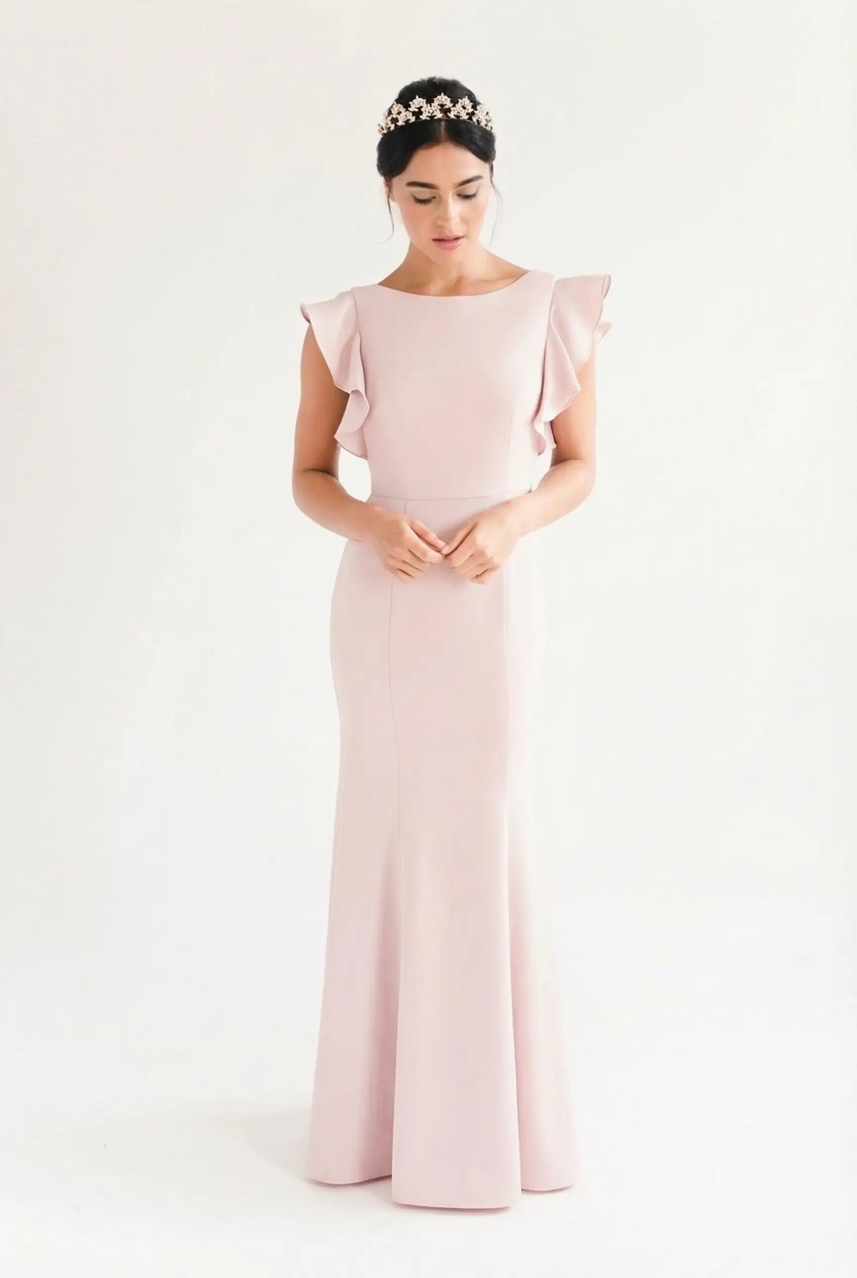 A woman in the TH&TH Cecelia Moss Crepe Bridesmaid Dress in Smoked Blush, featuring ruffled sleeves, stands against a white background. Her dark hair is styled up with a floral headband, and she gazes down with hands gently clasped.