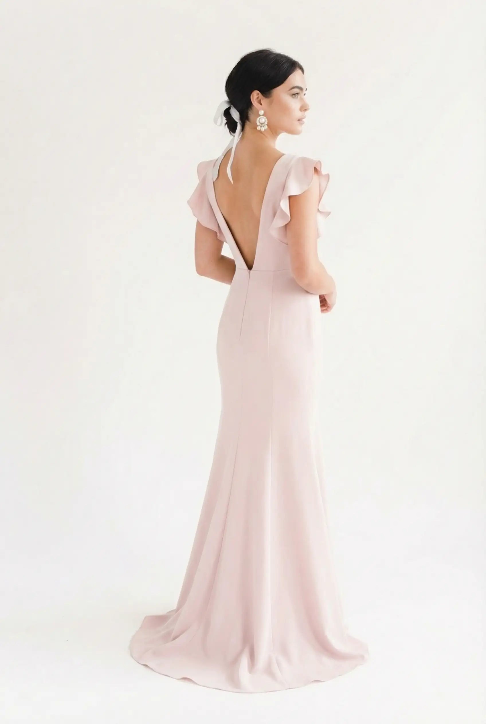 A woman with dark hair in a low bun and statement earrings wears the TH&TH Cecelia Moss Crepe Bridesmaid Dress in Smoked Blush—floor-length, featuring a deep V-back, ruffled sleeves, and halter tie—against a white background.