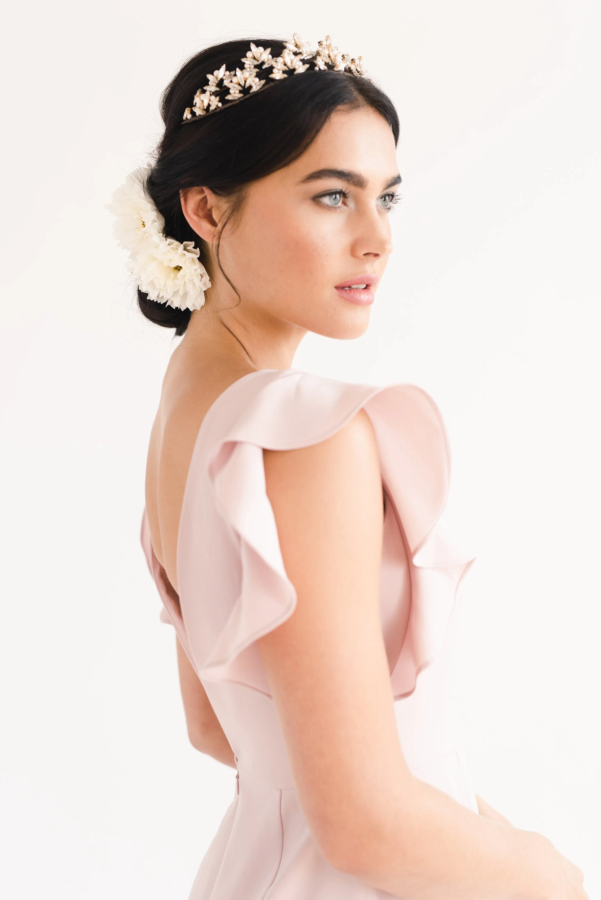A woman wearing the TH&TH Cecelia Moss Crepe Bridesmaid Dress in Smoked Blush, a size-inclusive gown with ruffled sleeves, looks to the side. Her dark hair is styled in an elegant updo with white flowers and a decorative floral headband.