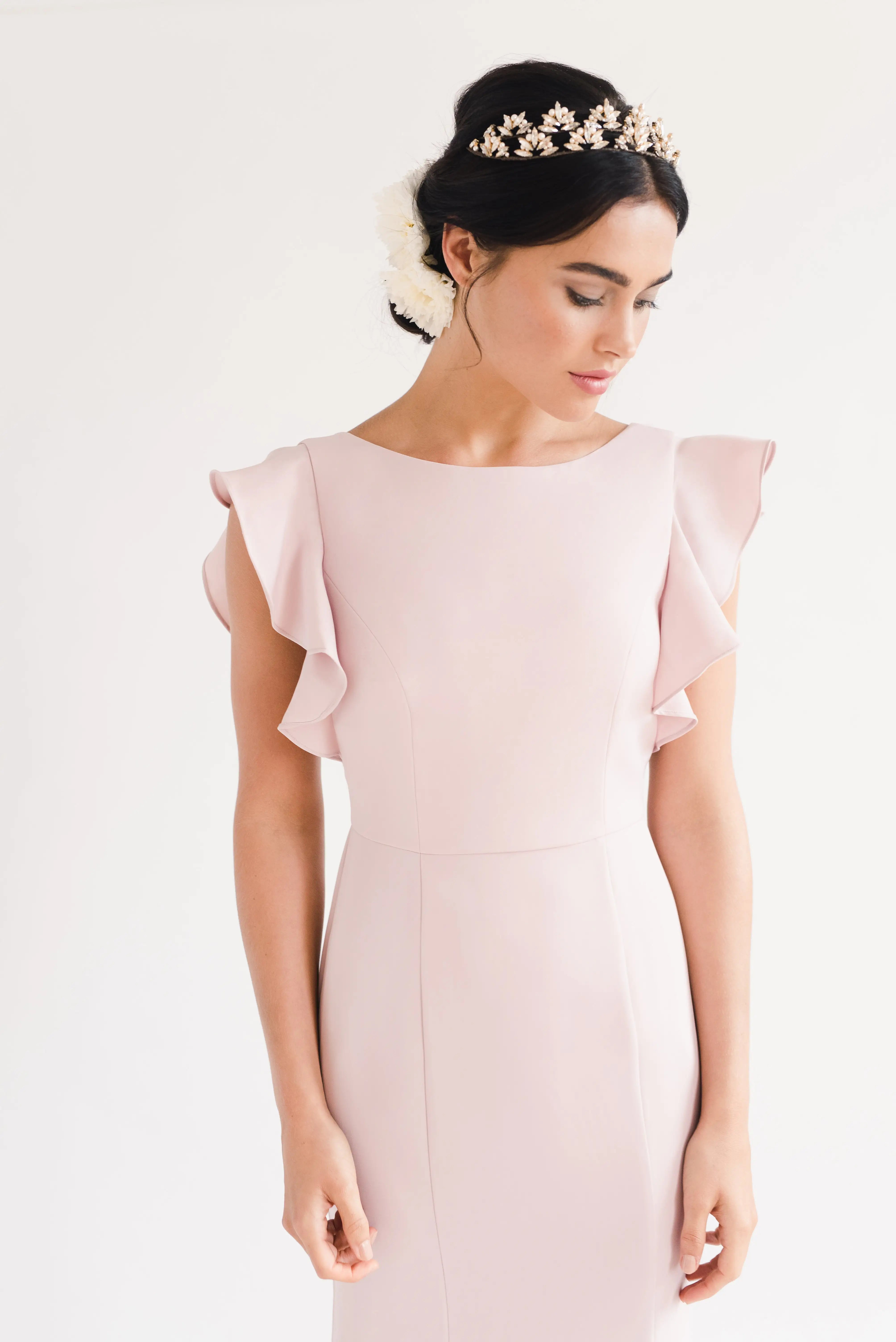 A woman in the TH&TH Cecelia Moss Crepe Bridesmaid Dress in Smoked Blush, a size-inclusive gown with ruffled shoulders, stands against a white background. She wears a floral headpiece and her dark hair is styled in an updo.