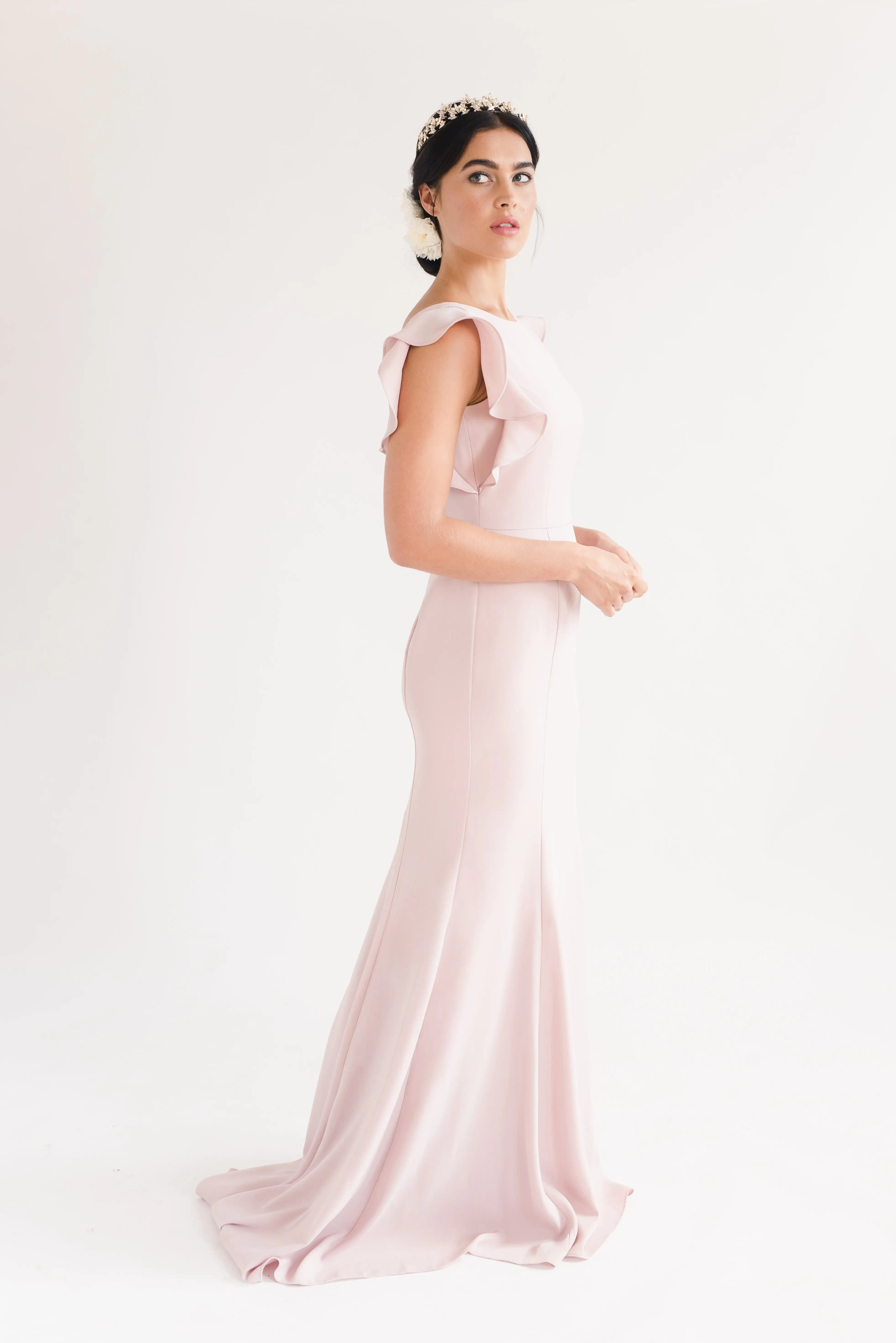A woman wears the TH&TH Cecelia Moss Crepe Bridesmaid Dress in Smoked Blush, featuring ruffled sleeves, against a white background. Her dark hair is styled up with a decorative headband as she gazes slightly to the side.