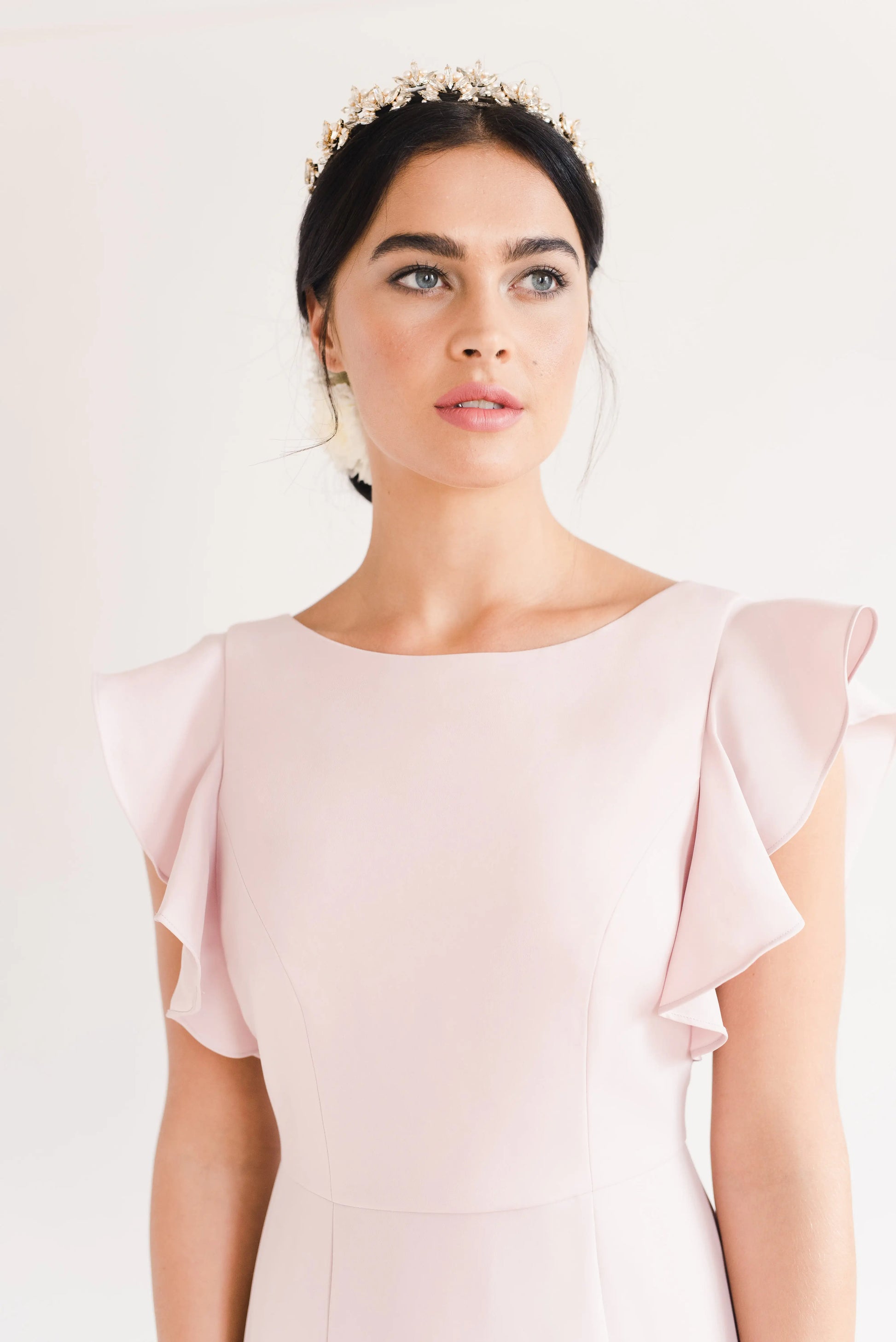 A woman with dark hair pulled back, wearing the TH&TH Cecelia Moss Crepe Bridesmaid Dress in Smoked Blush and a decorative headband, gazes upward against a plain light background. The gown features ruffled sleeves and a size-inclusive fit.