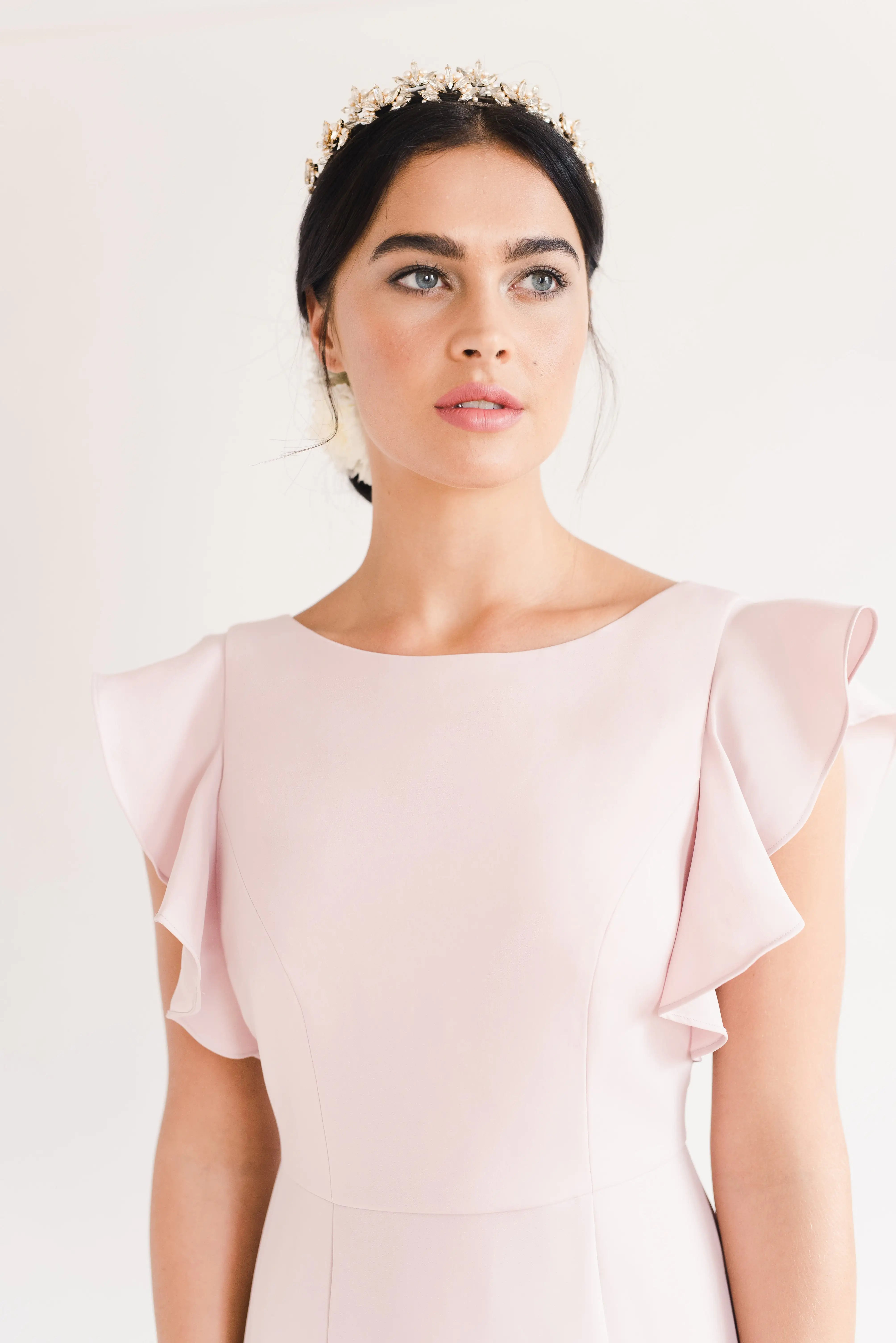 A woman with dark hair pulled back, wearing the TH&TH Cecelia Moss Crepe Bridesmaid Dress in Smoked Blush and a decorative headband, gazes upward against a plain light background. The gown features ruffled sleeves and a size-inclusive fit.