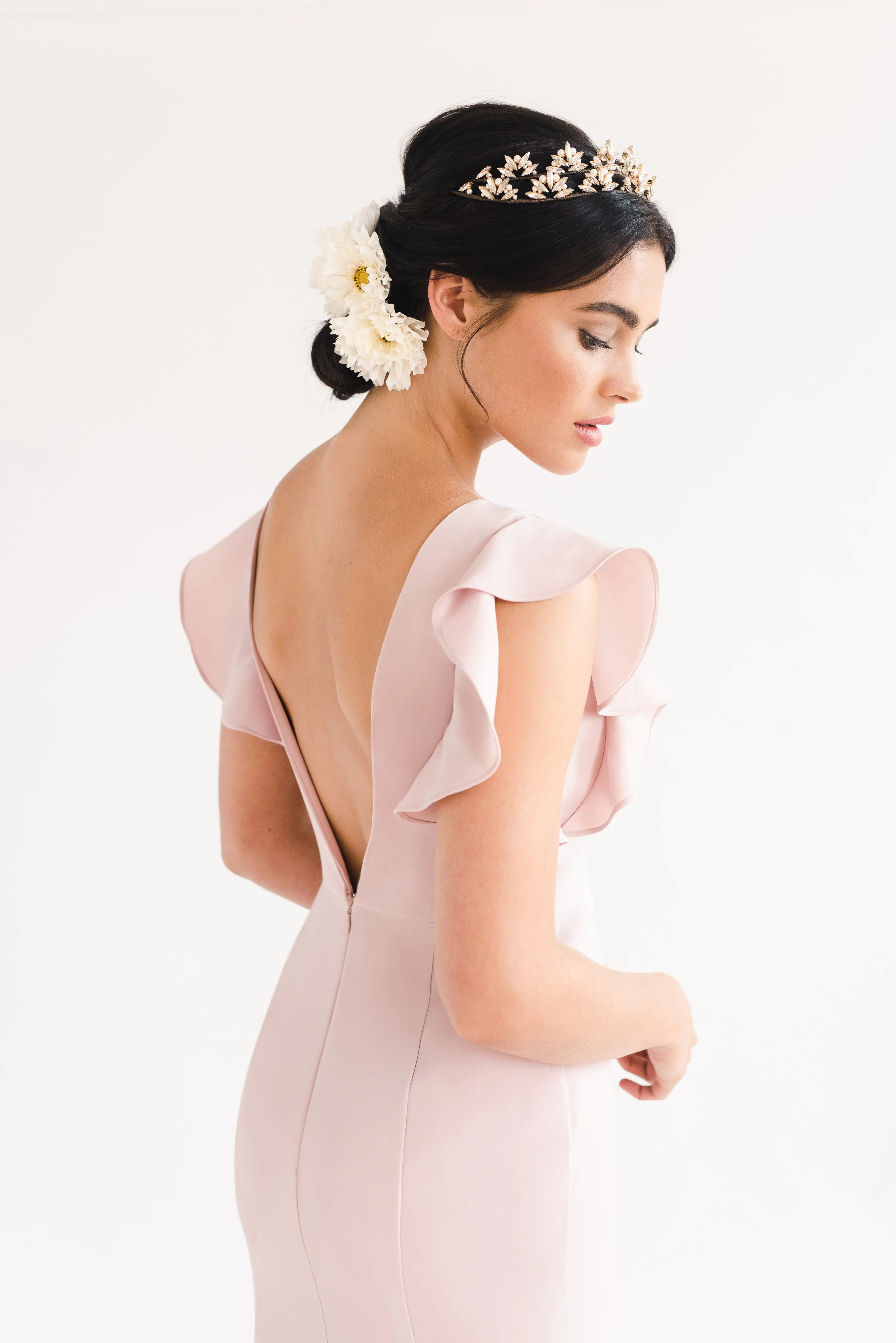 A woman in profile wears the TH&TH Cecelia Moss Crepe Bridesmaid Dress in Smoked Blush, featuring flutter sleeves and an open back. Her dark hair is styled in a low updo with a floral hairpiece and gold tiara, set against a plain white background.